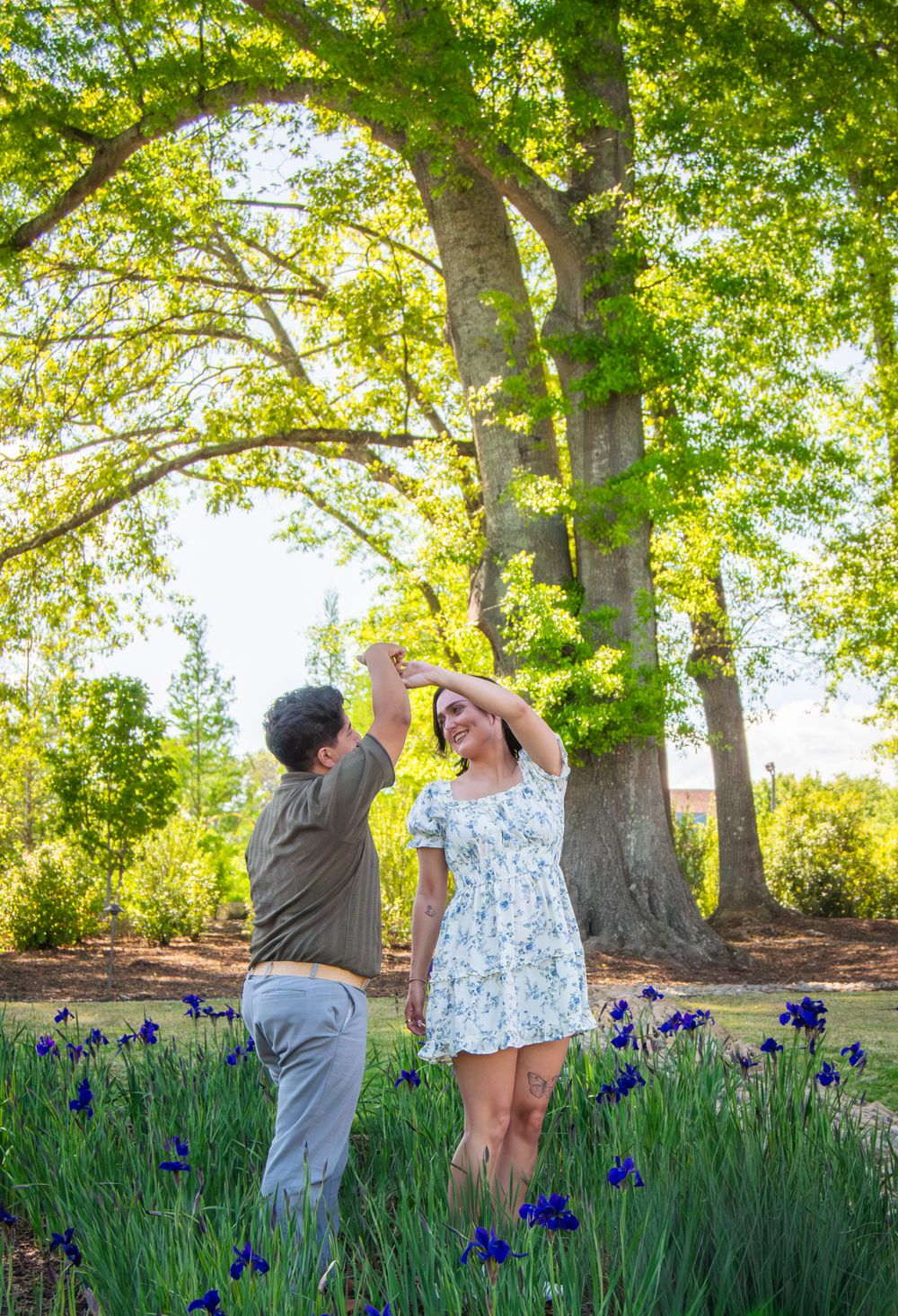 Couple dancing outdoors near blooming purple irises under a large tree in a sunny green park.
