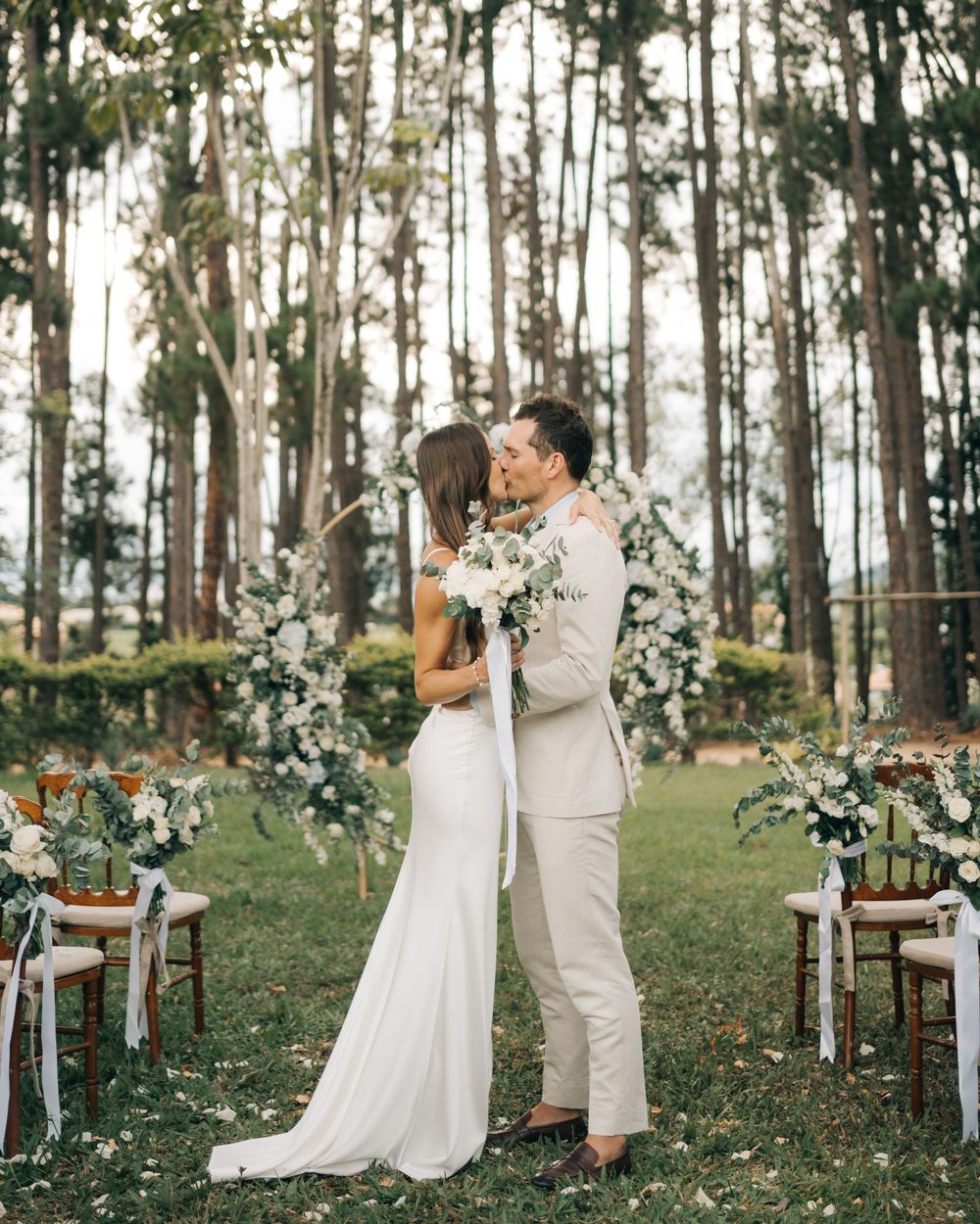 noivos se beijando em cerimônia de casamento ao ar livre no bosque com decoração romântica e convidados ao fundo