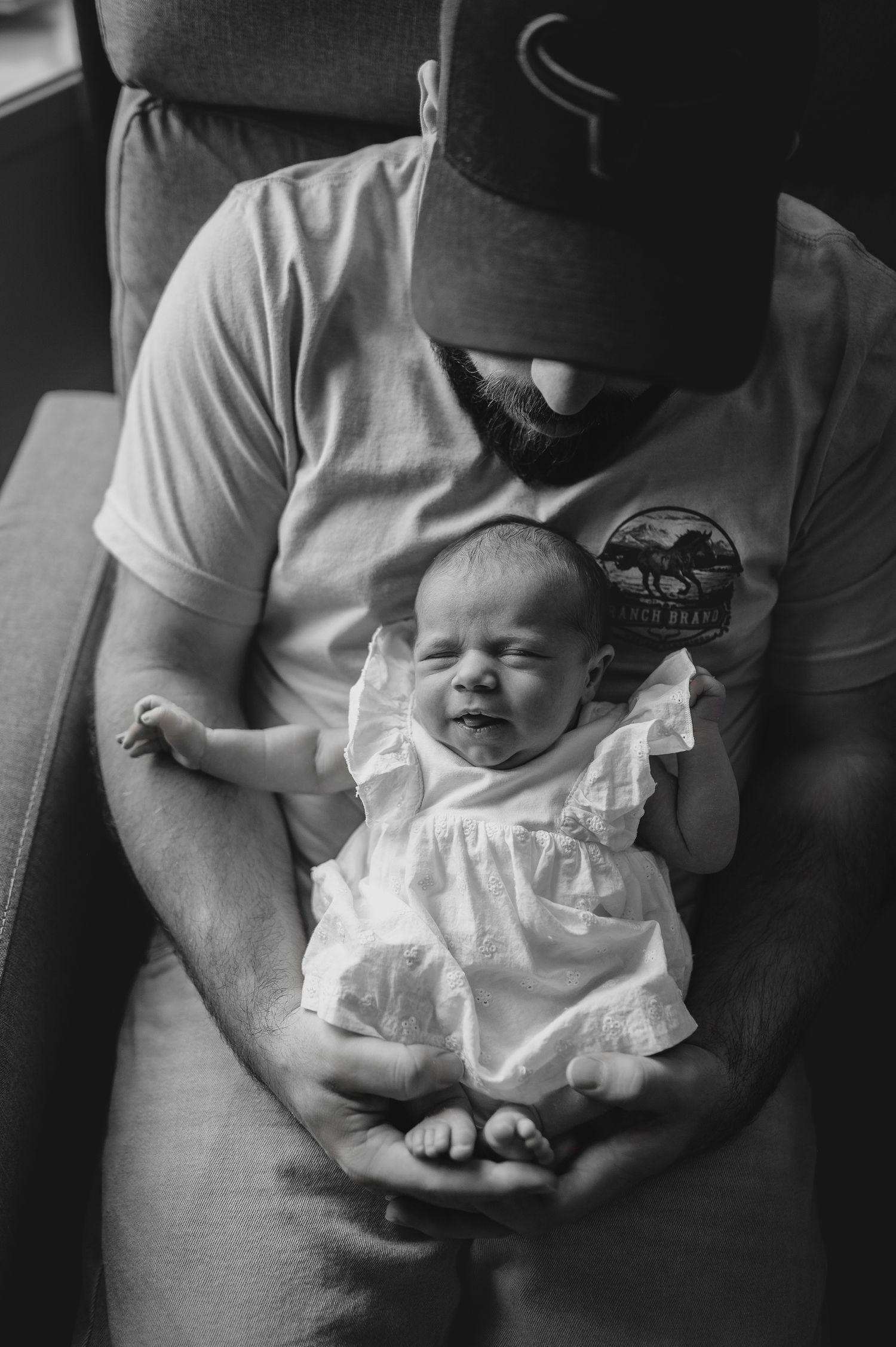 A tender black and white portrait shows a baby in a white dress resting peacefully in gentle arms.