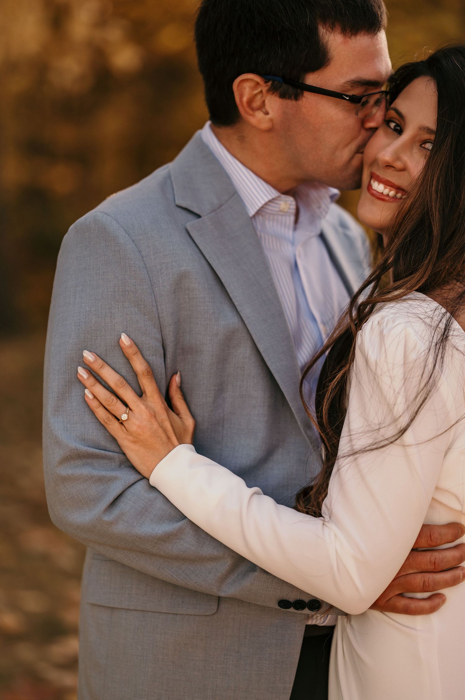 A romantic engagement photo showing a couple embracing outdoors in autumn light, with her displaying a ring.