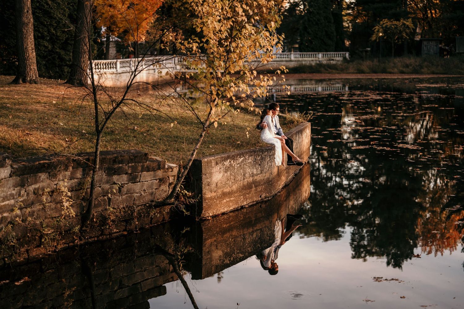 A couple stands near a tranquil pond surrounded by autumn trees with golden foliage reflecting in the water.