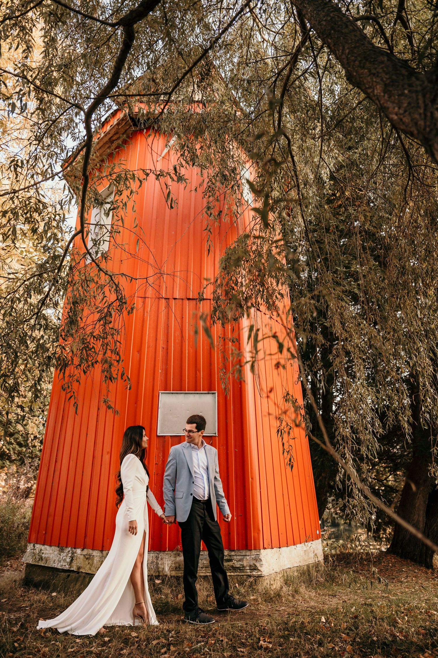 A couple poses in front of a vibrant red barn wall surrounded by autumn trees during a wedding photo session.
