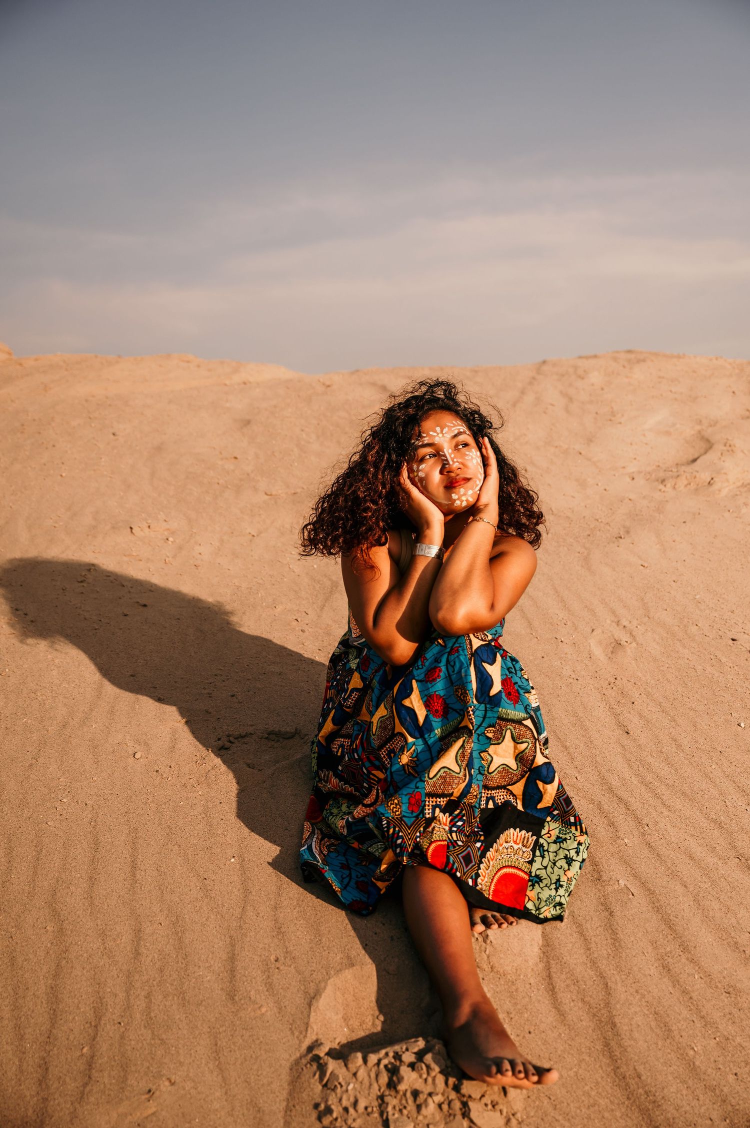 A person in a colorful patterned dress poses expressively on sandy dunes at sunset.