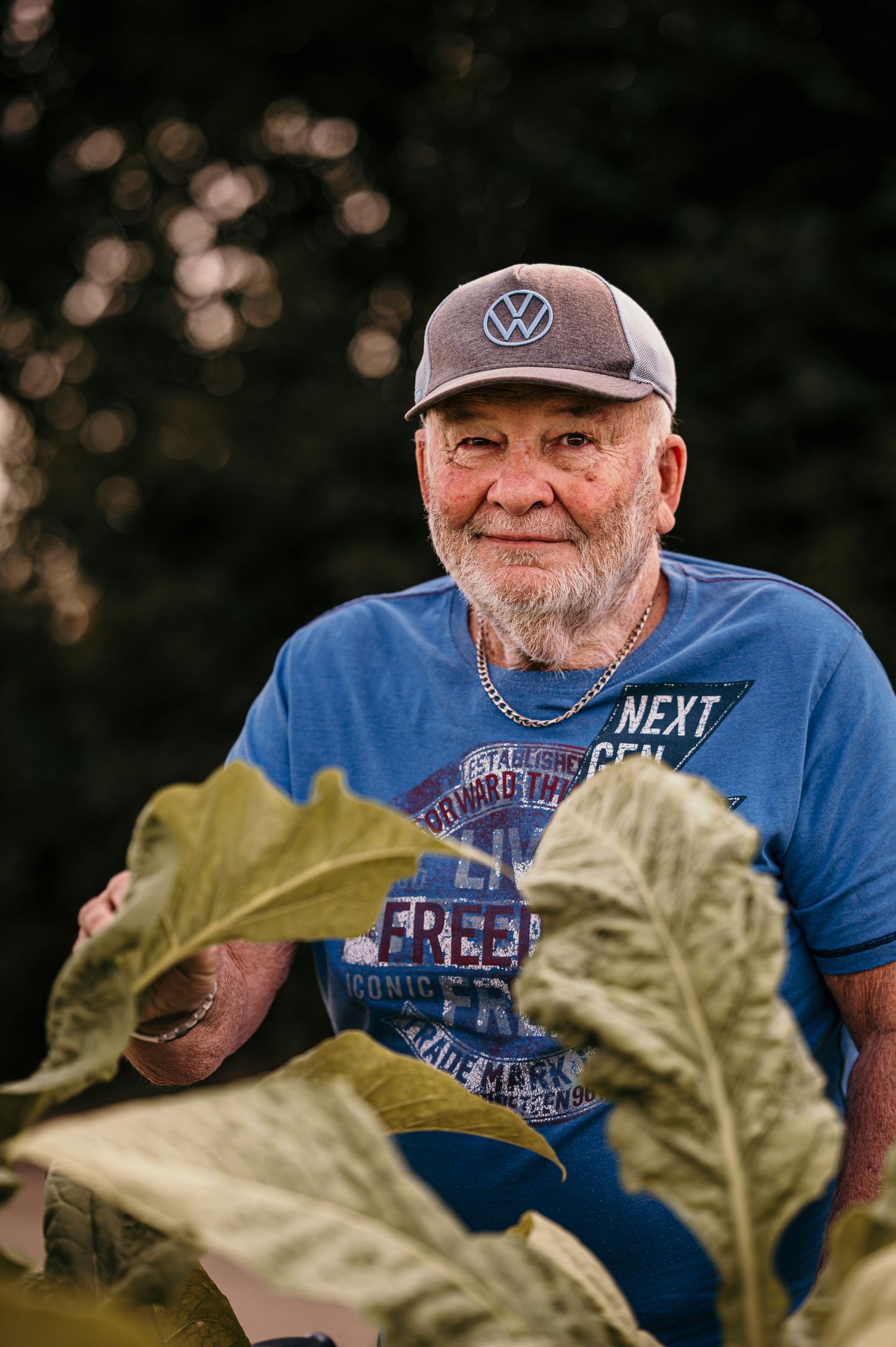 A person wearing a blue t-shirt and baseball cap smiles warmly behind large green leaves in an outdoor setting.