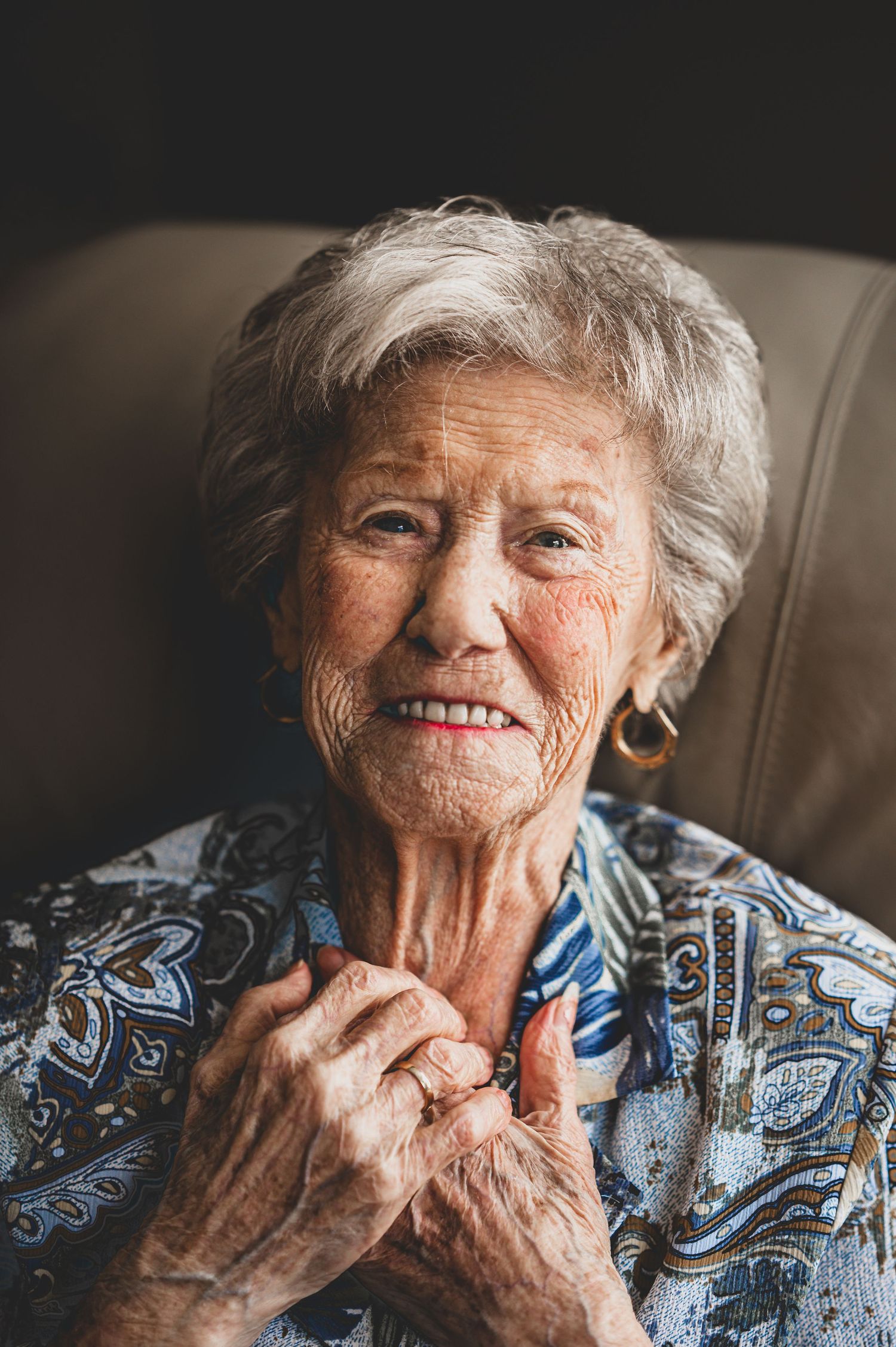 An elderly person wearing a blue and white patterned top smiles warmly while touching their face.