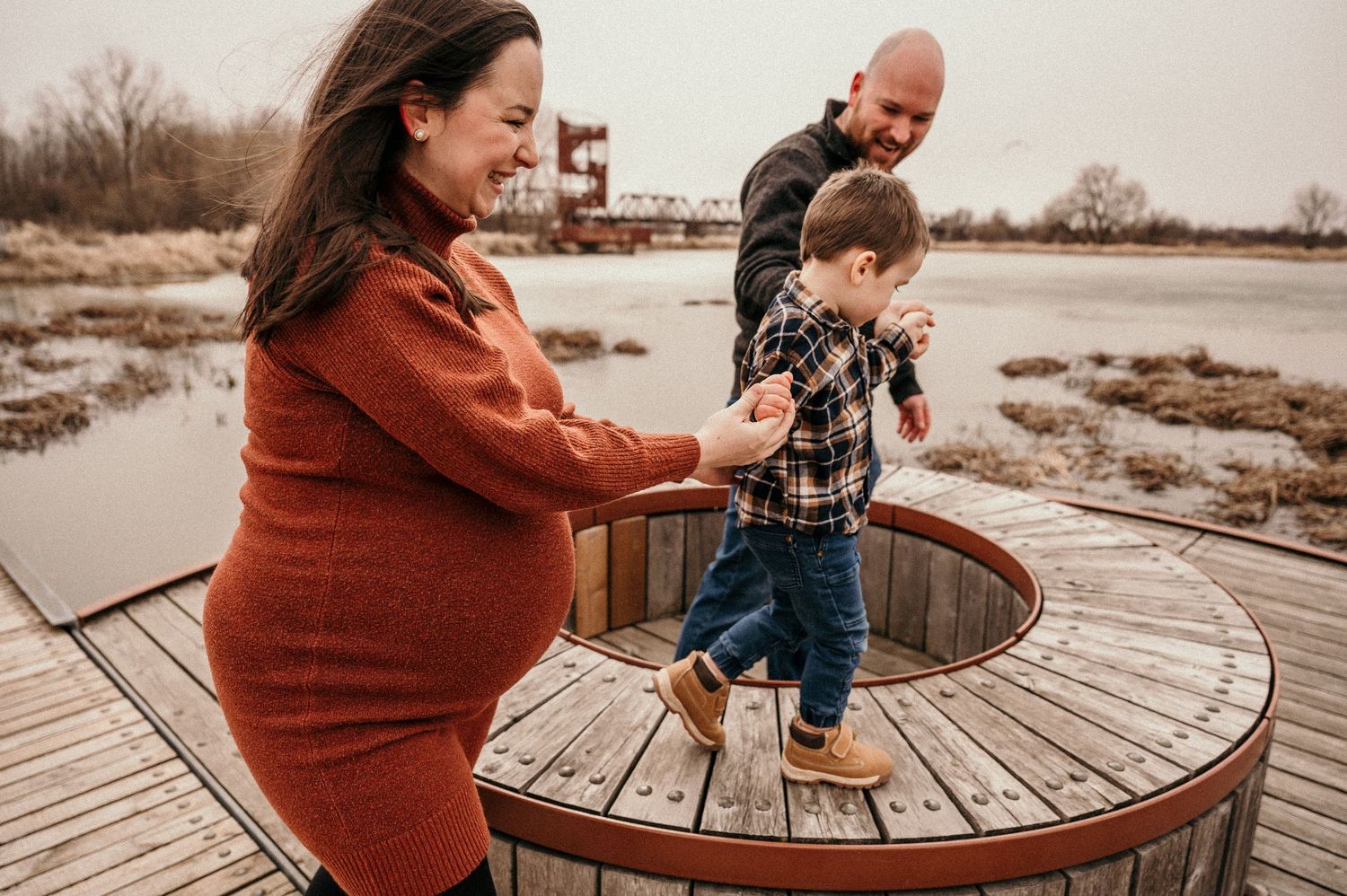 Pregnant woman in rust sweater holds toddler's hand on wooden dock while bald man watches near a serene lake.