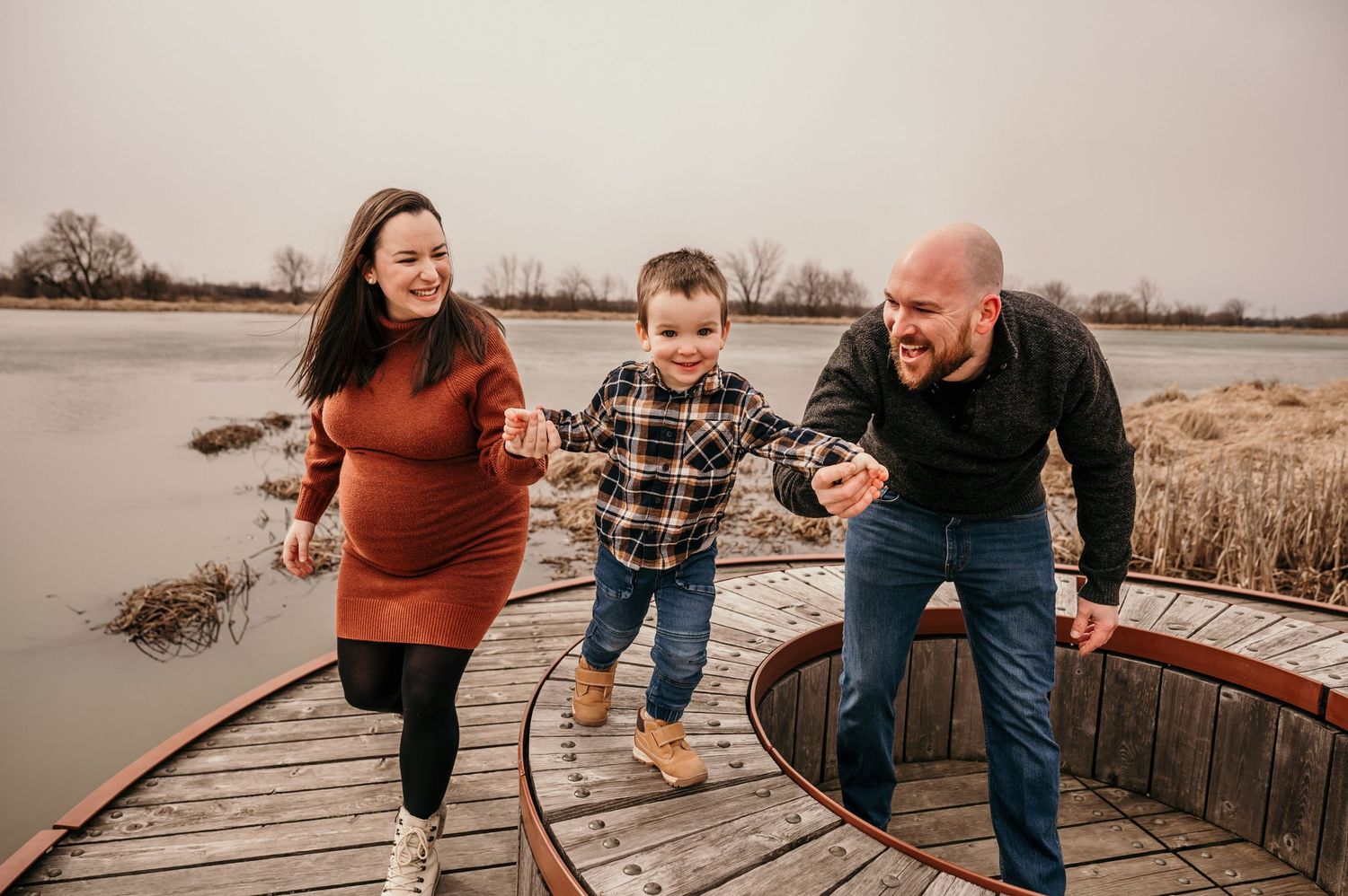 Happy family with pregnant mother, father, and young son playing on a wooden dock by a lake in autumn.