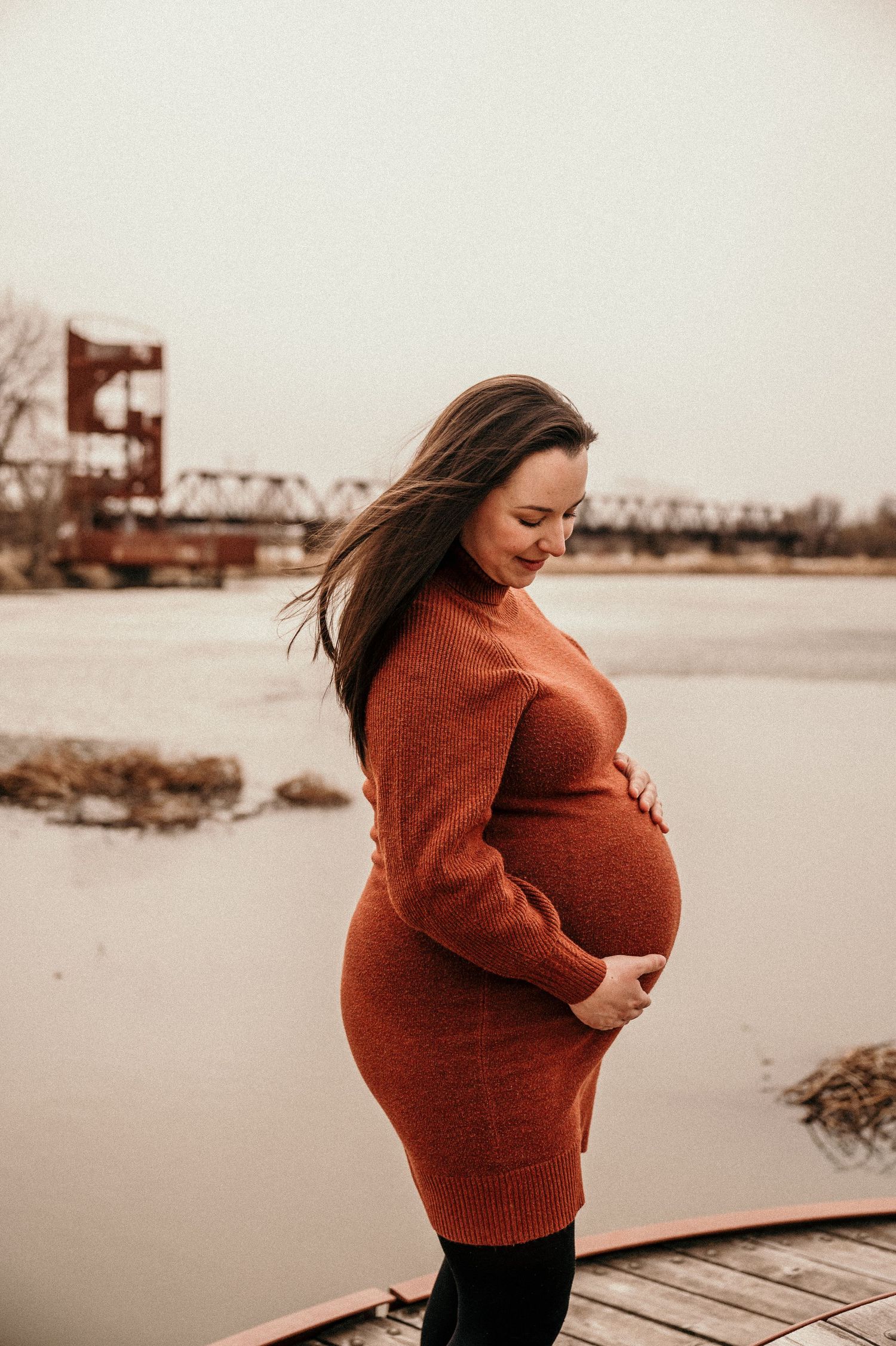 Pregnant woman in rust orange sweater dress cradling baby bump outdoors by a river with a bridge in the background.