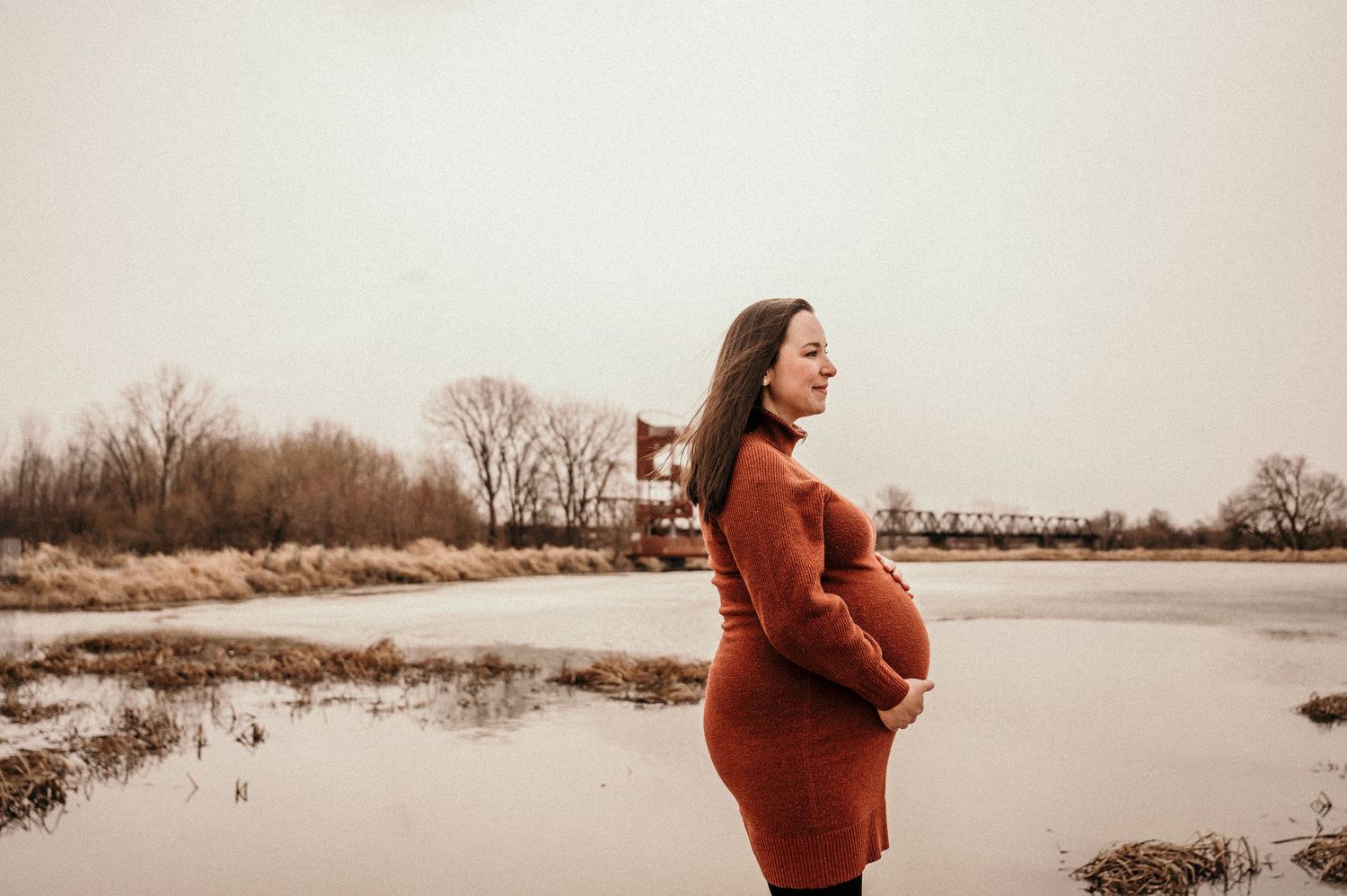 Pregnant woman in rust sweater dress cradling baby bump by a serene winter river landscape.