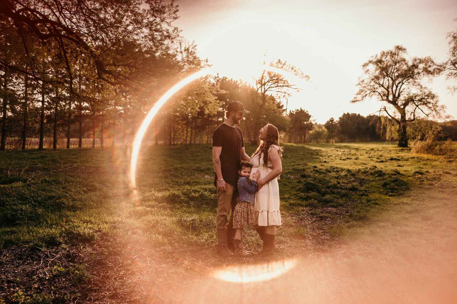 Family embracing in golden sunset light with circular lens flare in lush green park setting.