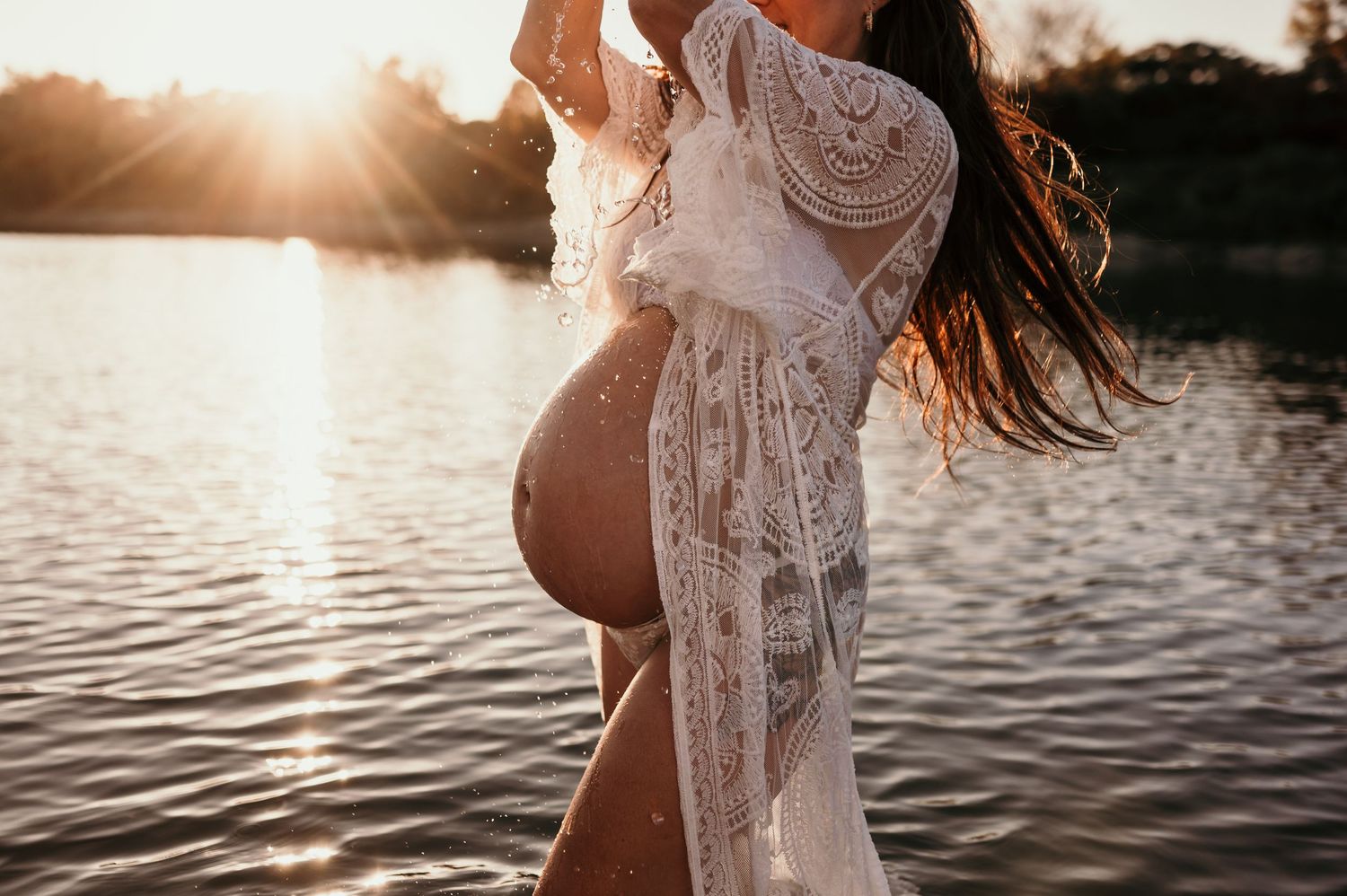 Silhouetted figure in a lace dress stands at the edge of a lake during golden hour sunset.