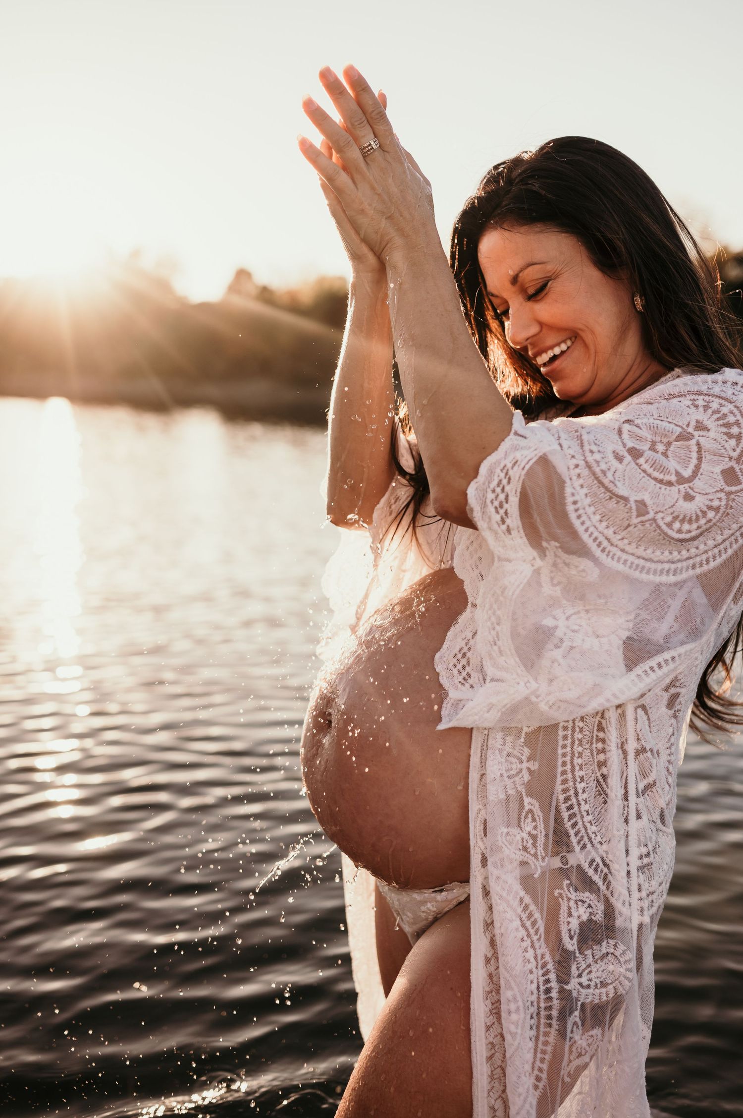 Maternity photoshoot at sunset shows woman in white lace dress splashing water at the beach.