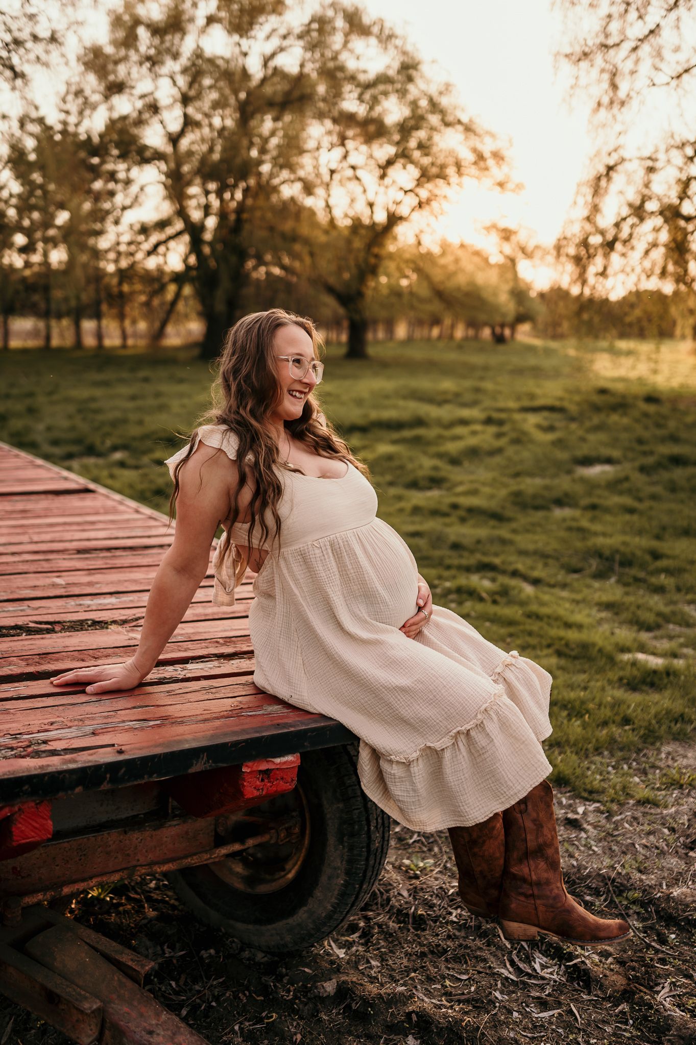 Pregnant woman in white dress and cowboy boots sits on rustic wooden trailer at golden hour on a farm.