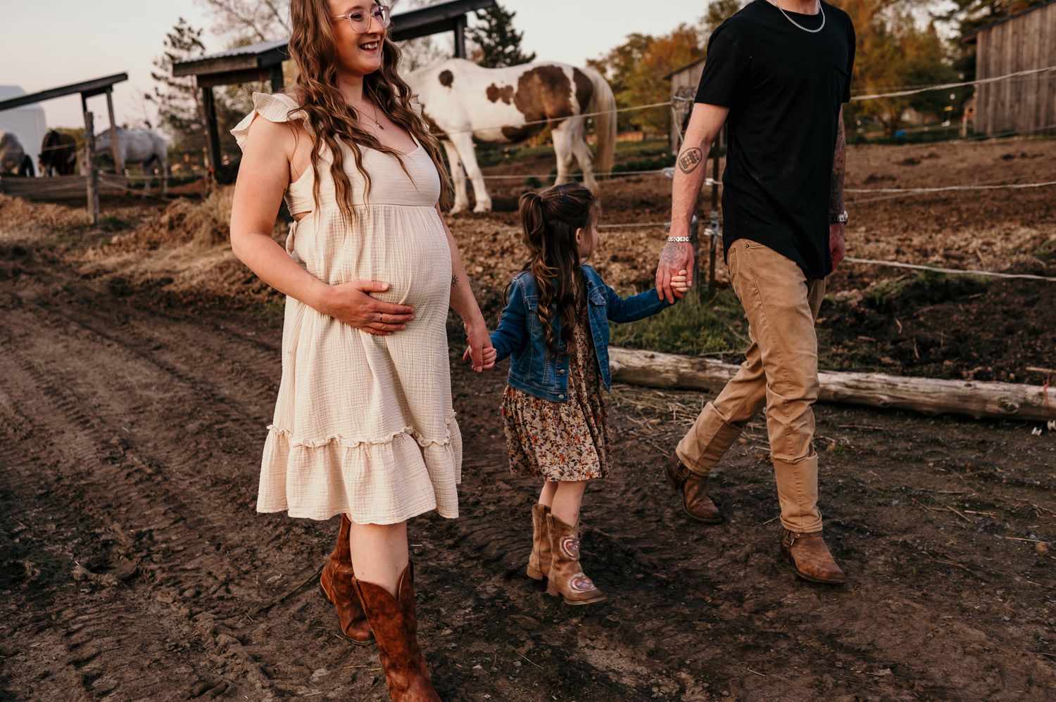 Pregnant woman in white dress walks with family on muddy farm path, holding hands with young girl, cow visible behind.