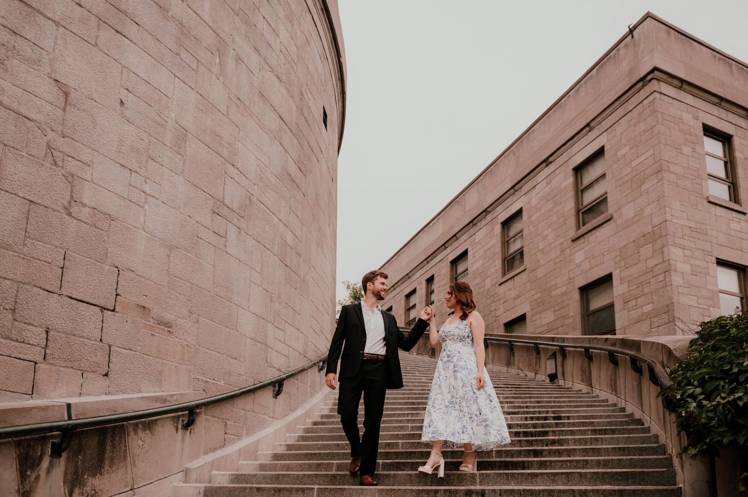 A couple ascends stone steps beside a historic building's curved wall.