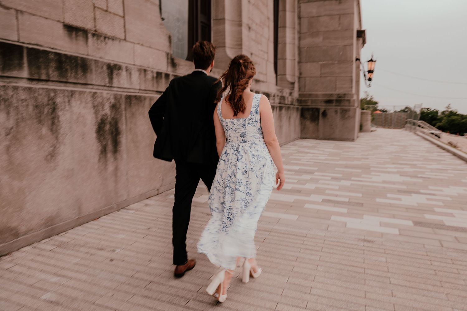 A couple walks along a historic stone building's terrace with the woman in a floral dress.