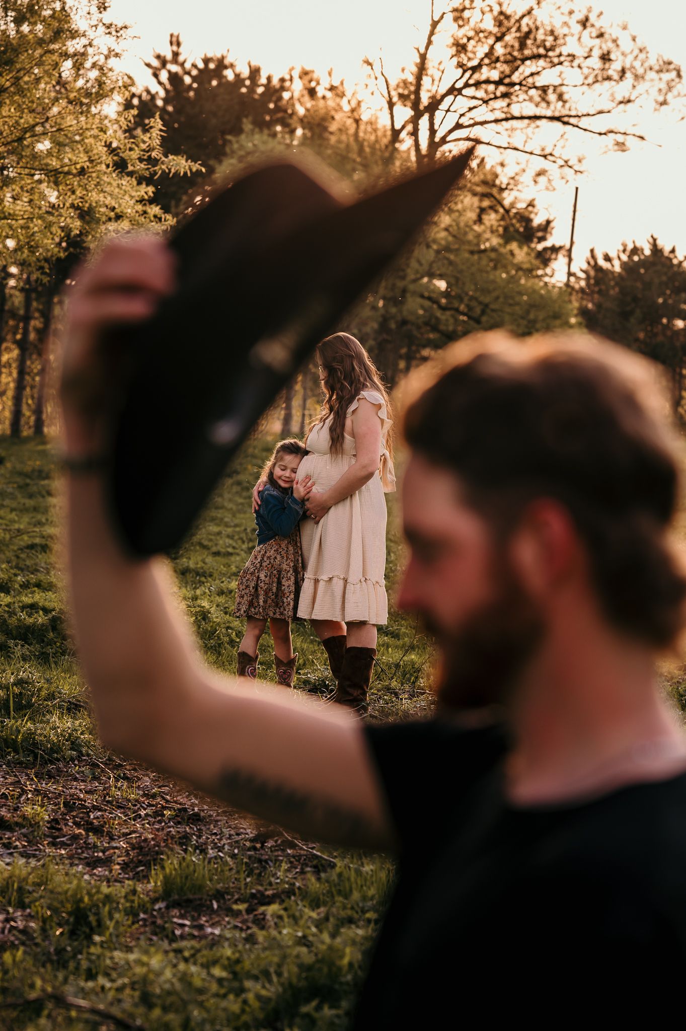A bearded man holds a cowboy hat while a woman in a cream dress embraces a child in a sunlit garden.