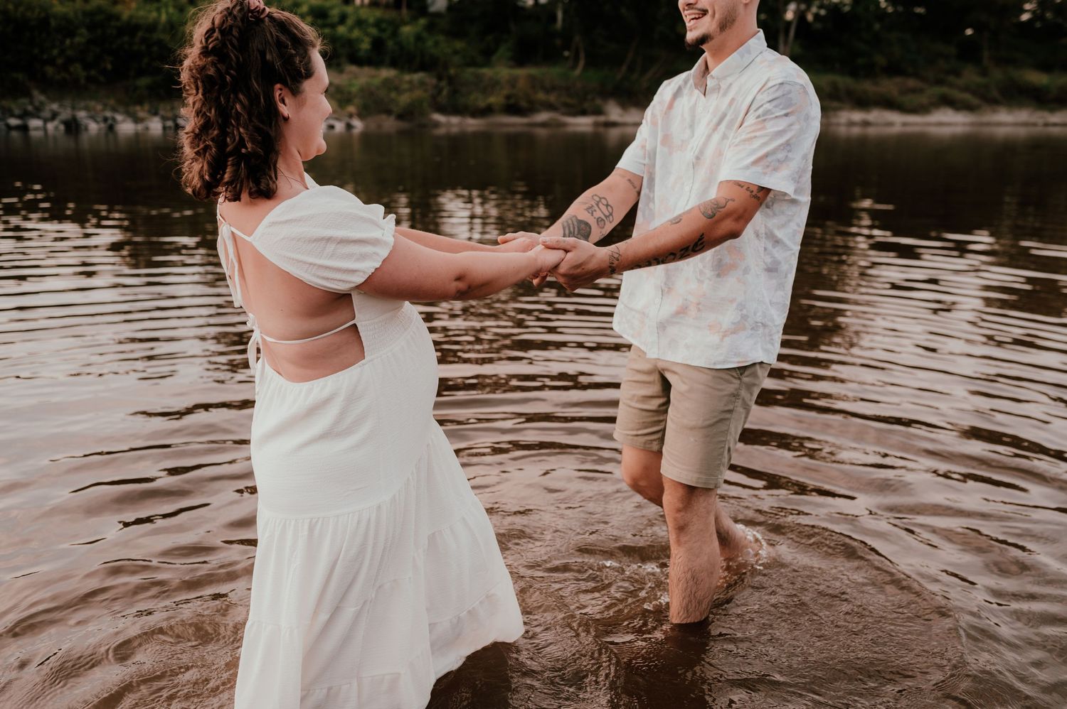 A couple holds hands while playing in shallow river water at sunset.