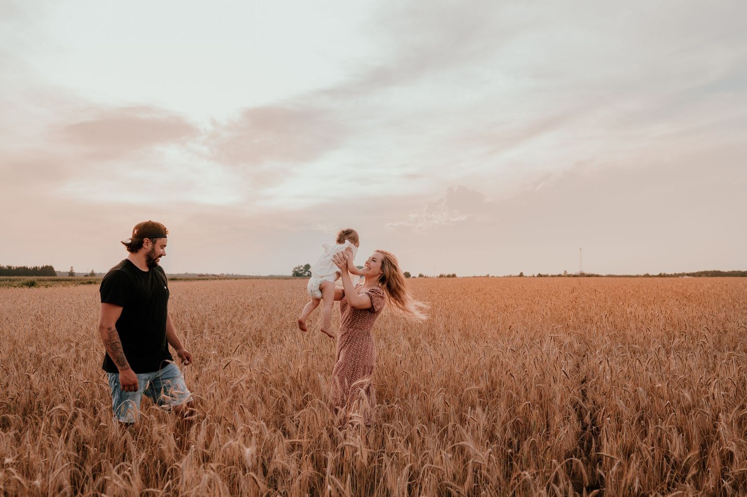 Three figures joyfully run through a golden wheat field at sunset with warm pink skies overhead.