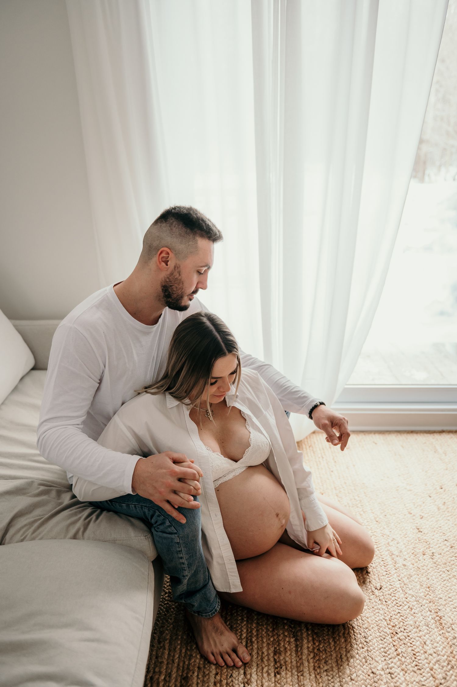 Intimate maternity photo series showing couple sitting together on carpeted floor by window in white room.