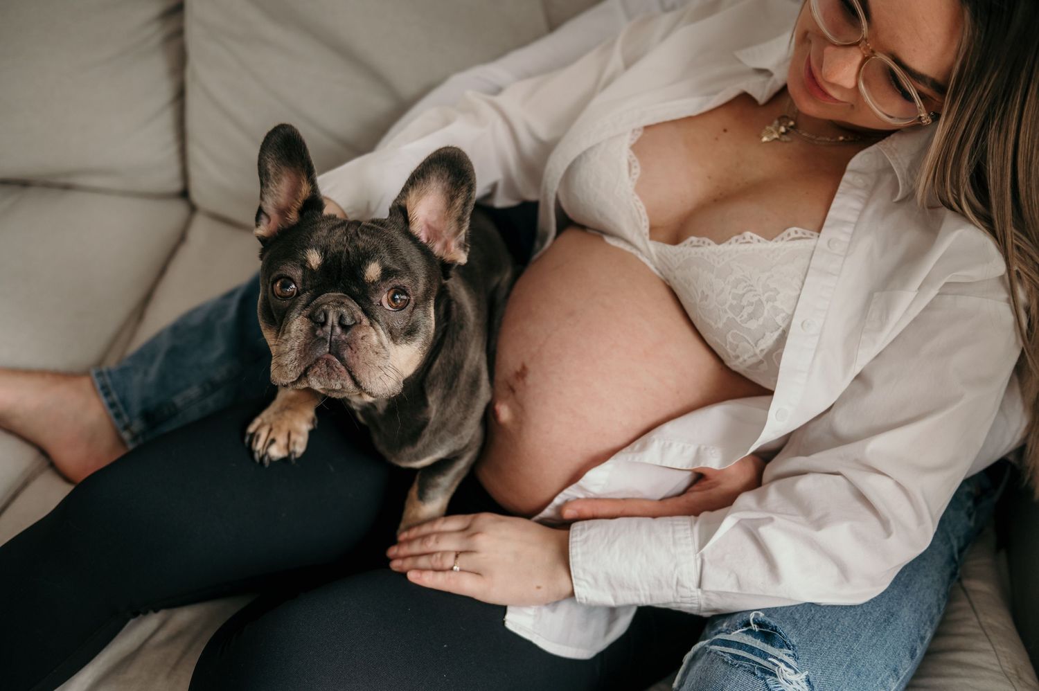 A French Bulldog rests beside an expectant mother on a couch during a maternity photoshoot.