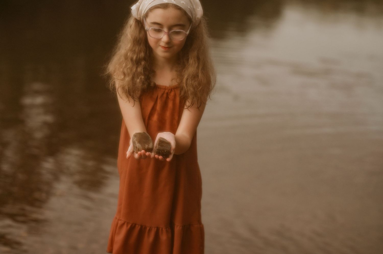 A young girl in a rust dress stands in shallow water, holding small stones in her outstretched hands.