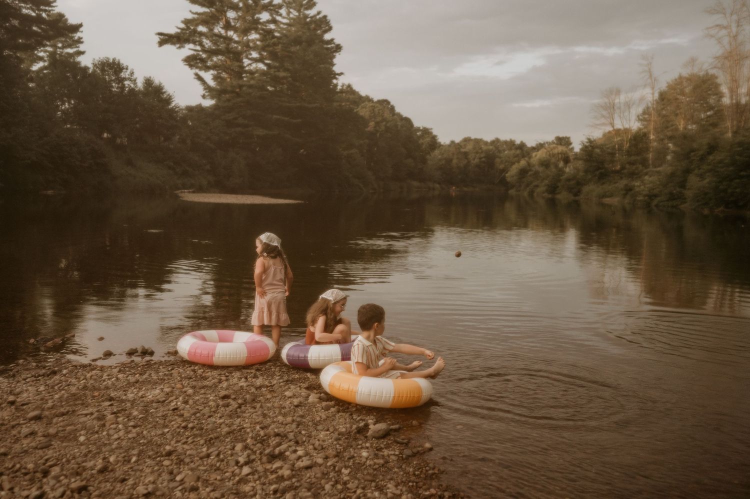 Children playing with colorful inflatable rings on a rocky riverbank surrounded by lush green trees.