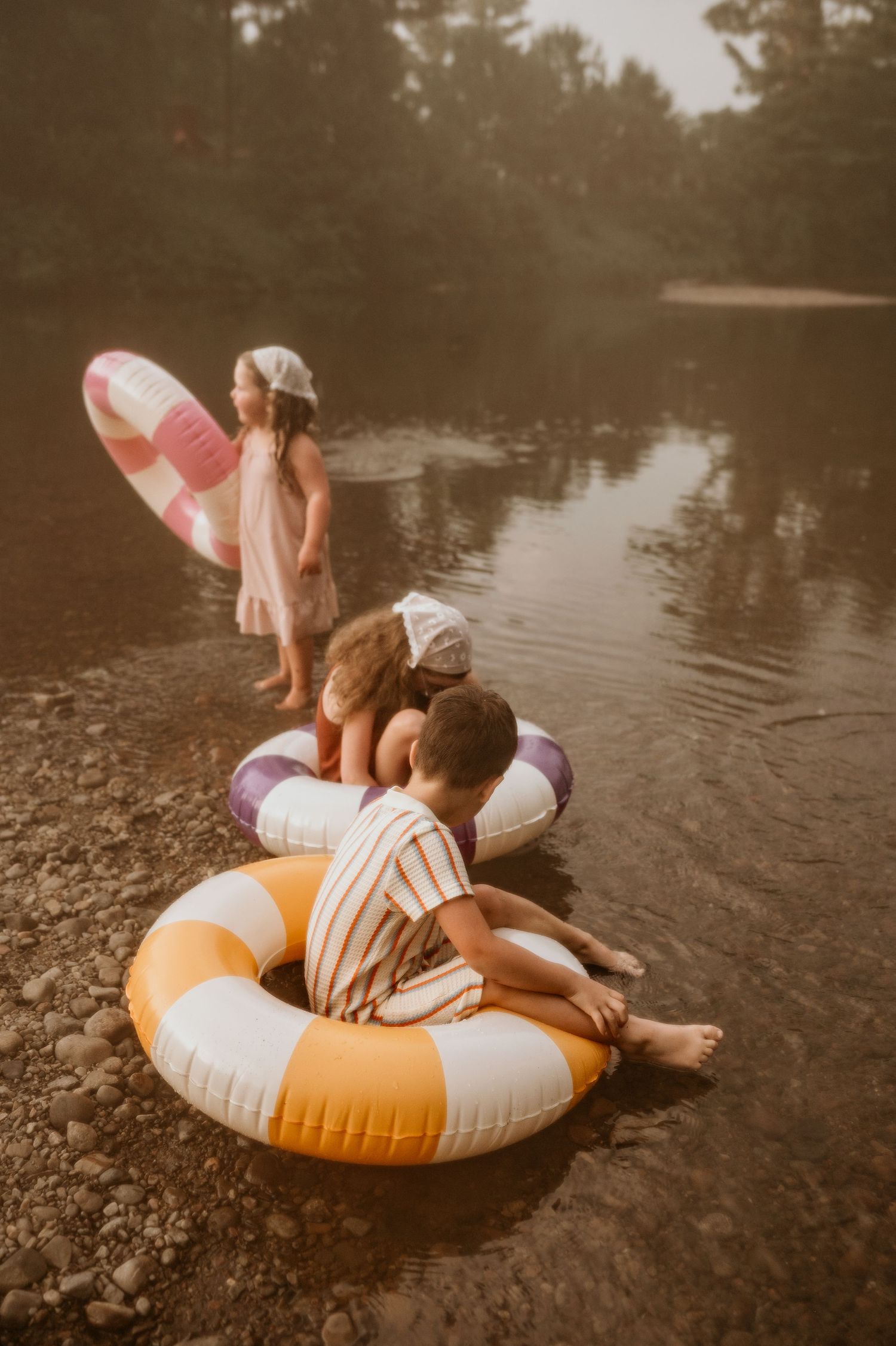 Children playing by a river with colorful inflatable pool rings on a misty summer day.