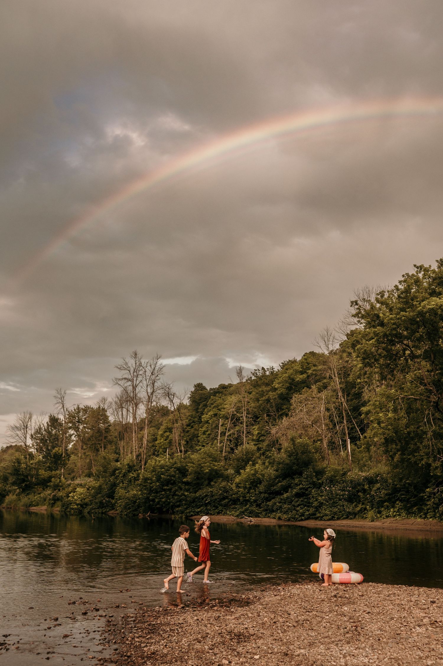 Family playing by a rocky riverbank under a stunning rainbow and dramatic cloudy sky, surrounded by lush green trees.
