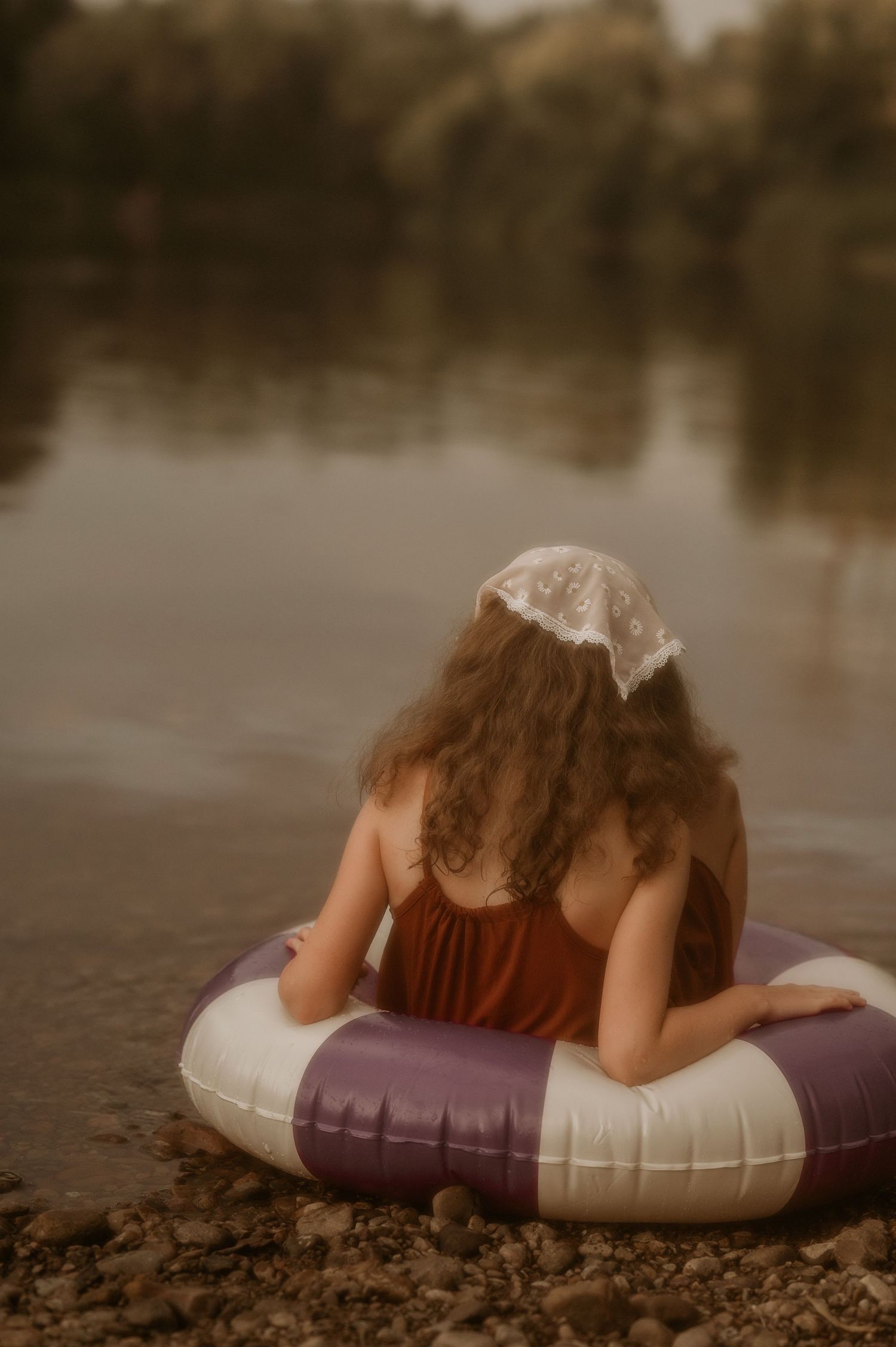 A young girl with curly hair sits by a calm lake, leaning on a purple inflatable ring, wearing a rust dress and lace bonnet.