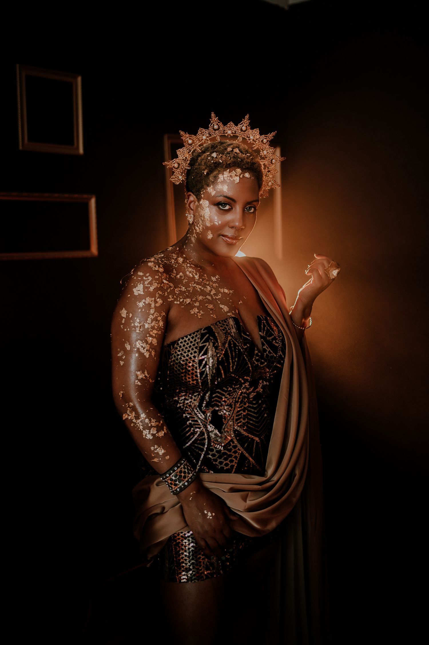 Figure in an ornate golden crown and embellished dress poses dramatically in moody lighting with glitter makeup effects.