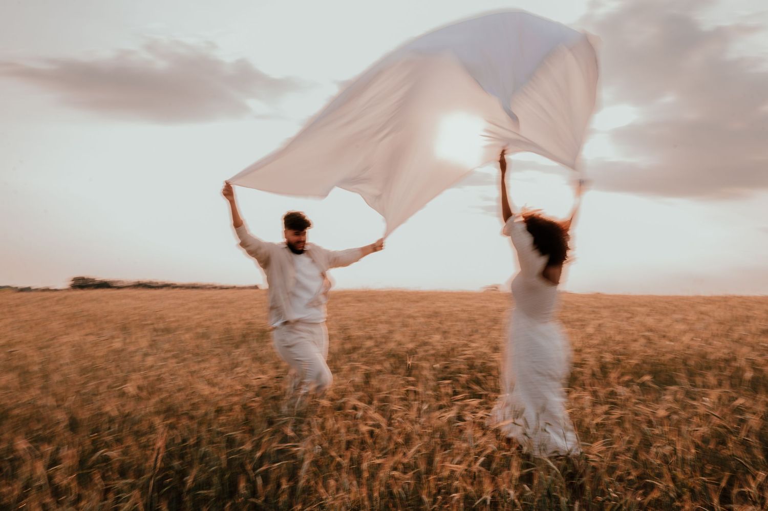 Two figures in white running through wheat field holding flowing fabric in the wind.