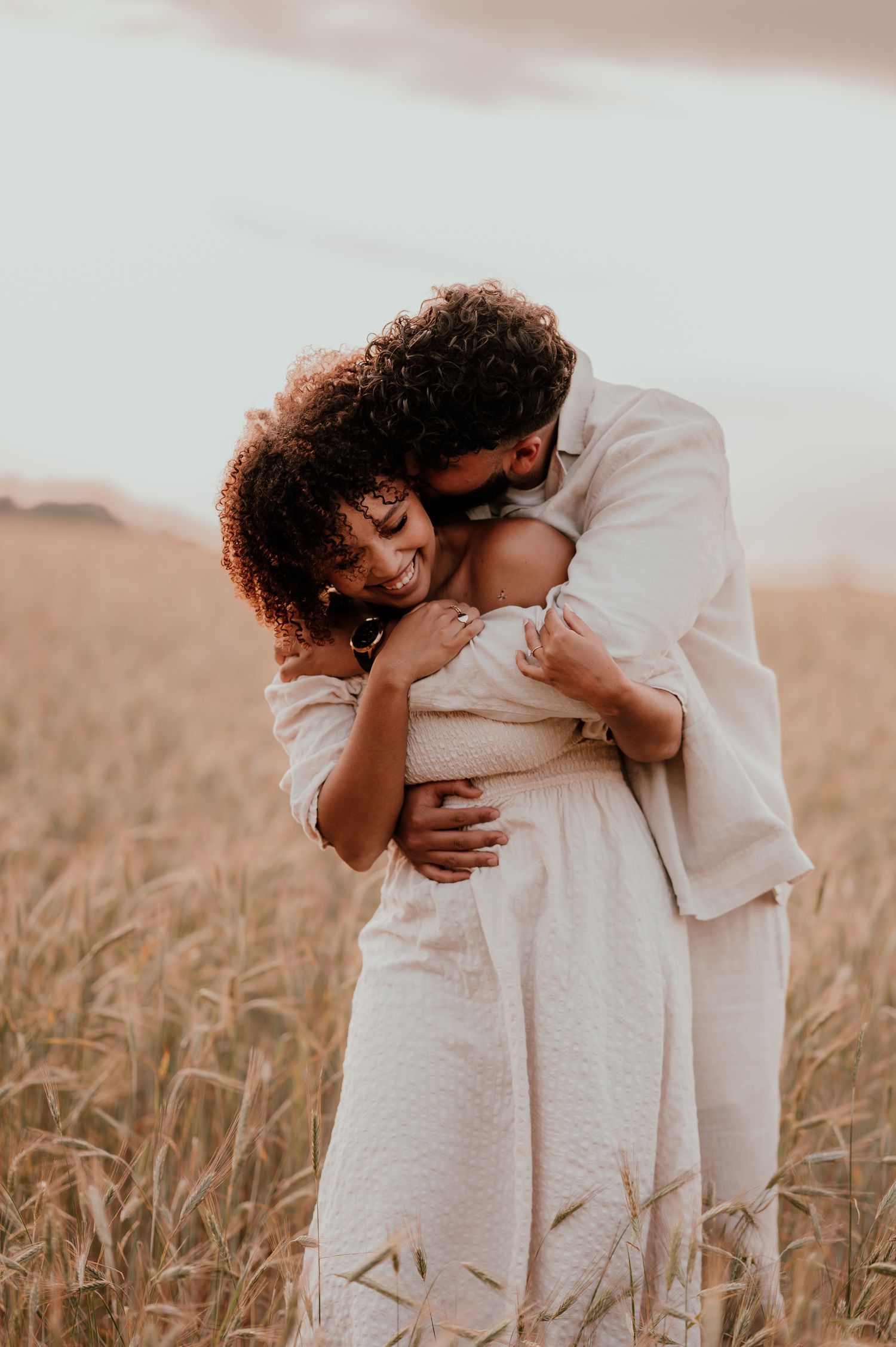 Romantic couple embracing in a golden wheat field at sunset wearing white outfits.