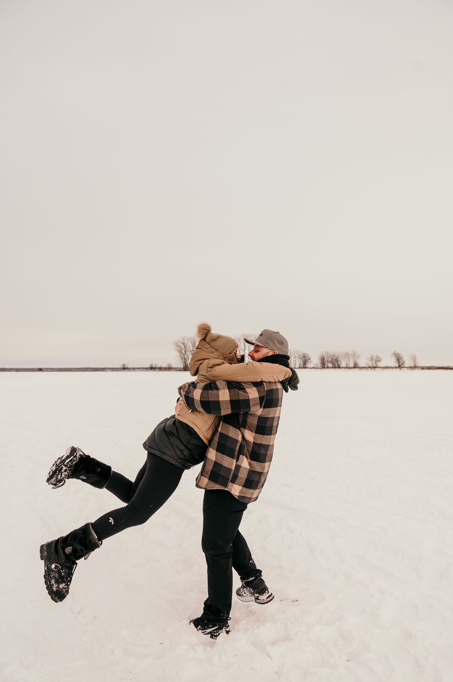 Two people embrace and spin together in a snowy winter field while wearing plaid and dark clothing.
