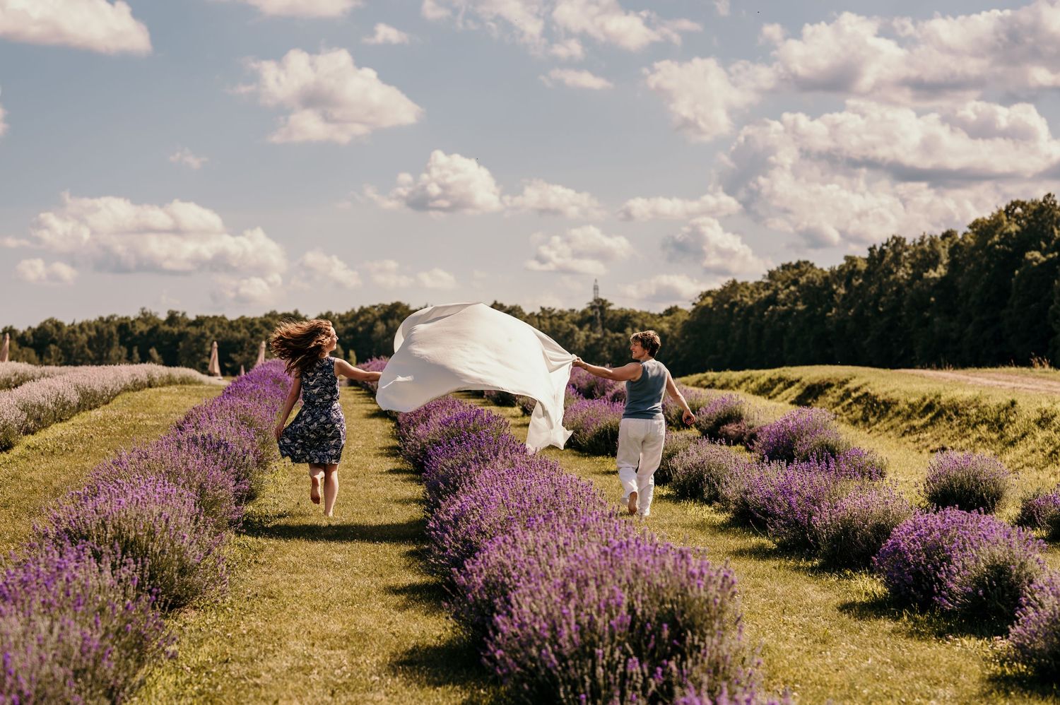Two women running through lavender fields holding a white fabric, with cloudy skies and trees in the background.
