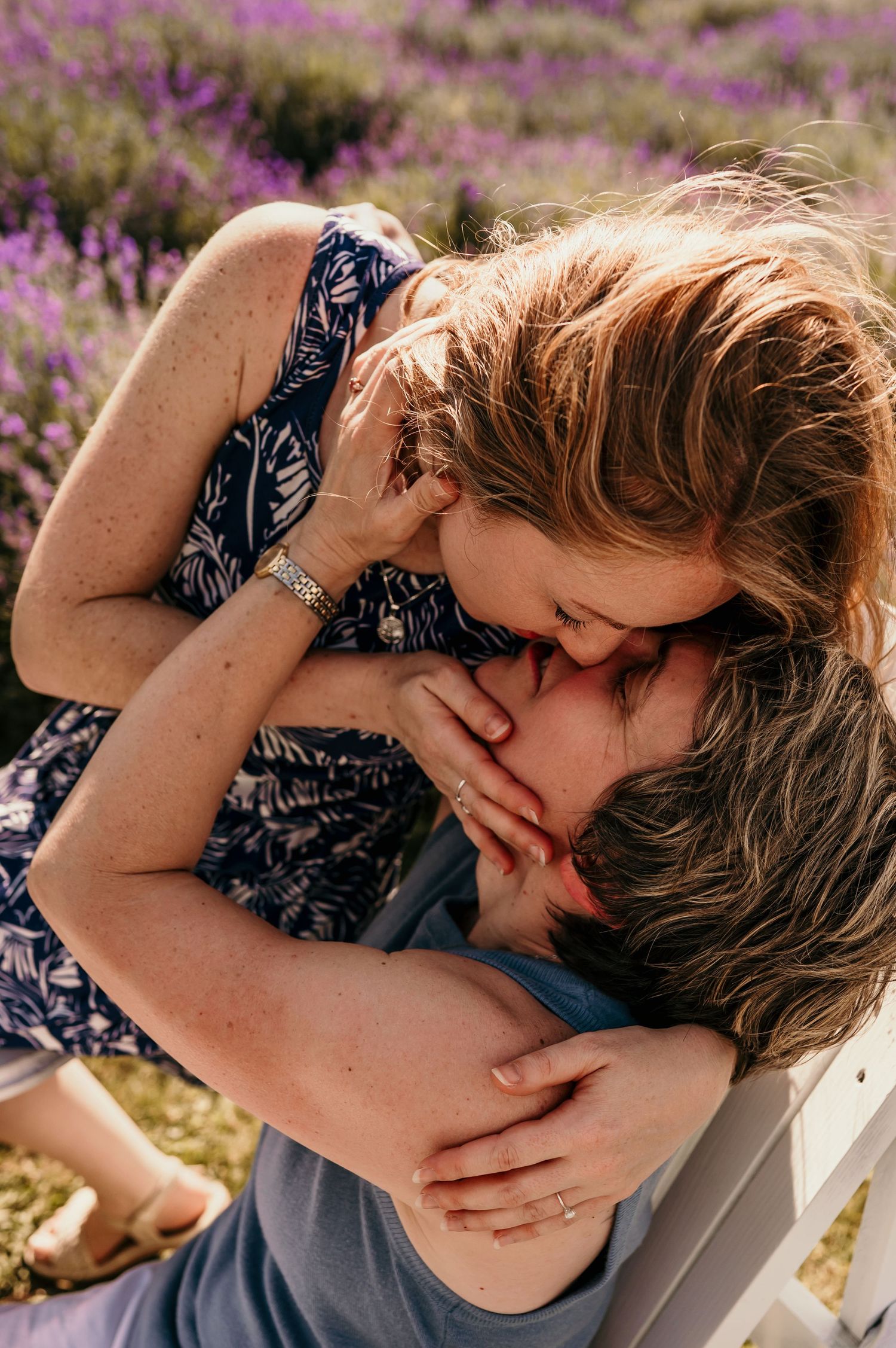 Couple sharing a romantic kiss in a blooming lavender field, woman in floral dress embracing man tenderly.