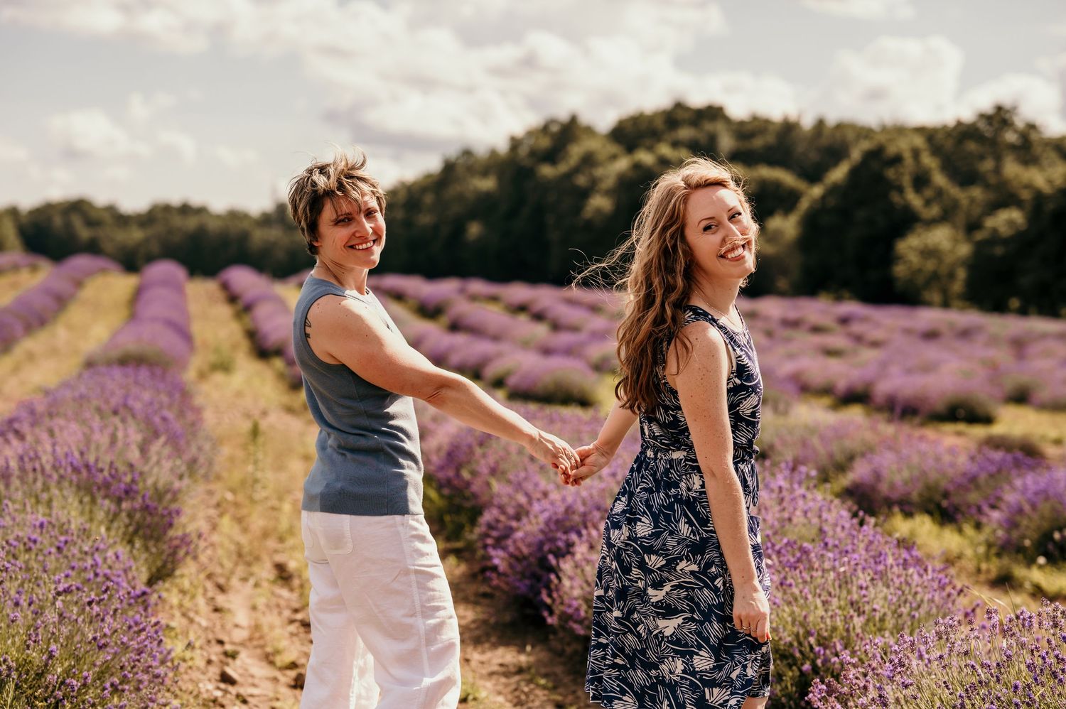 Two women holding hands and smiling in a vibrant purple lavender field on a sunny day.