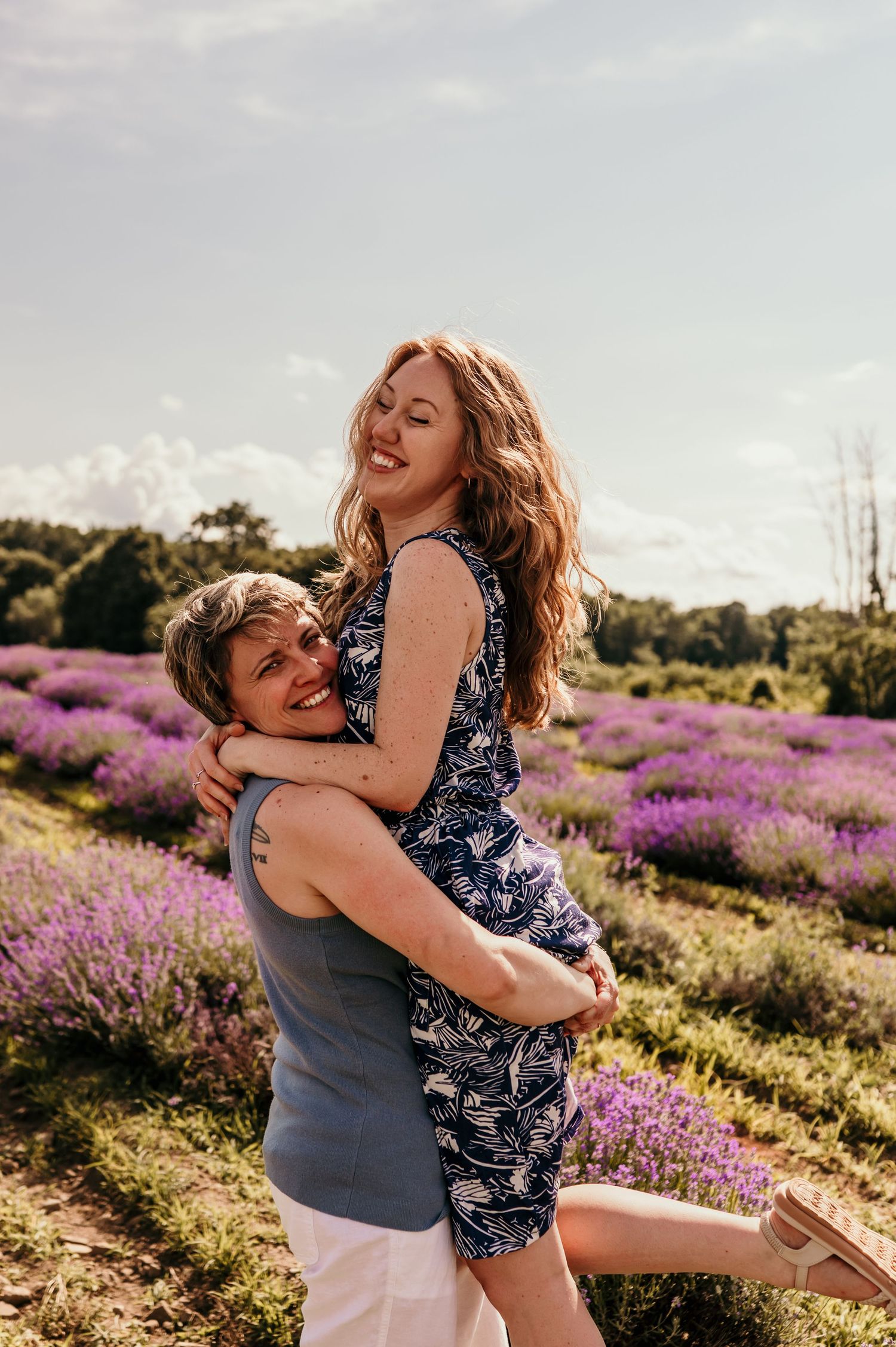 Couple embracing joyfully in a blooming lavender field on a sunny day, woman lifted by man in floral dress.