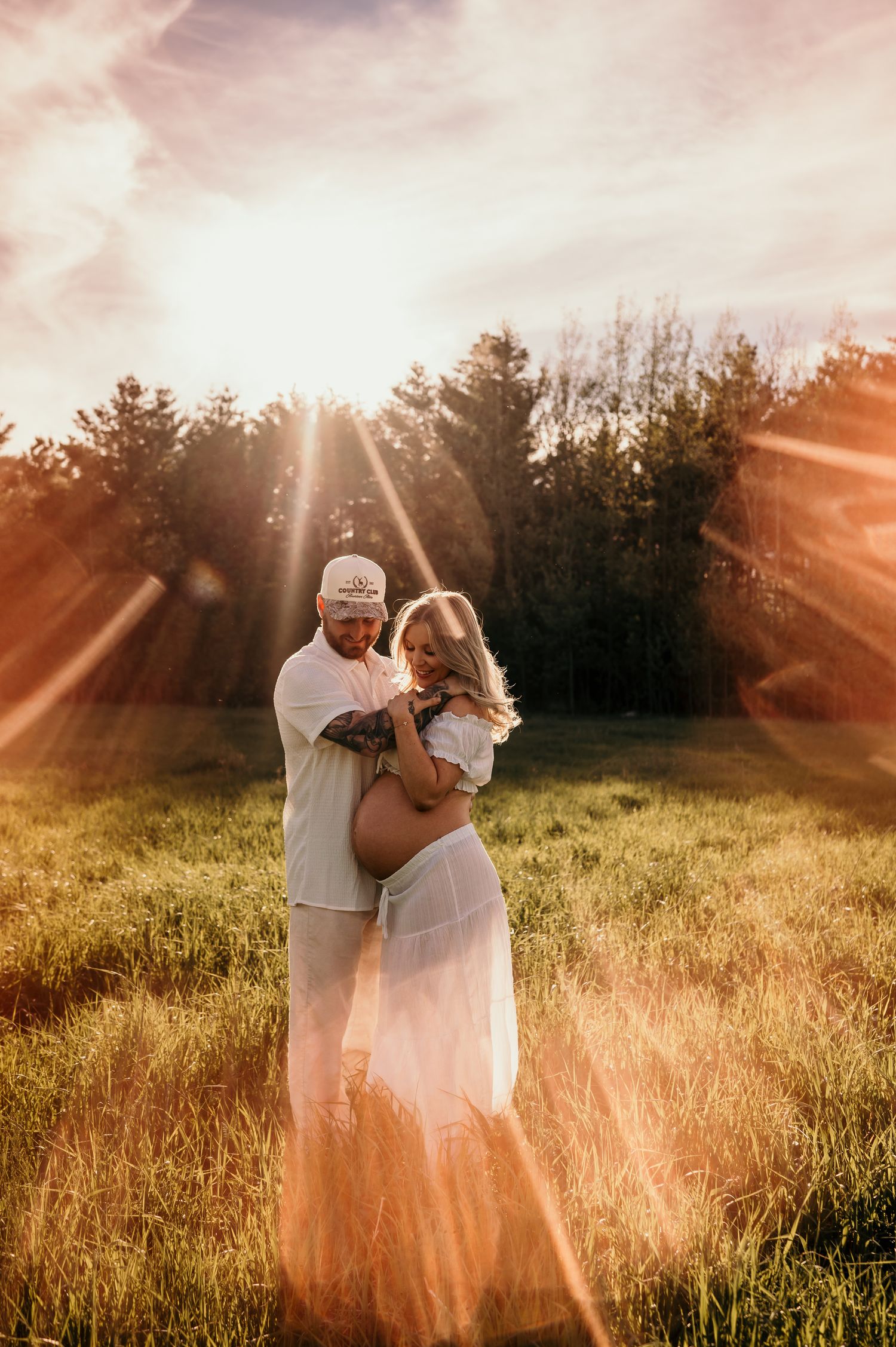 Couple embracing in sunlit field during golden hour maternity photoshoot with lens flare and trees.