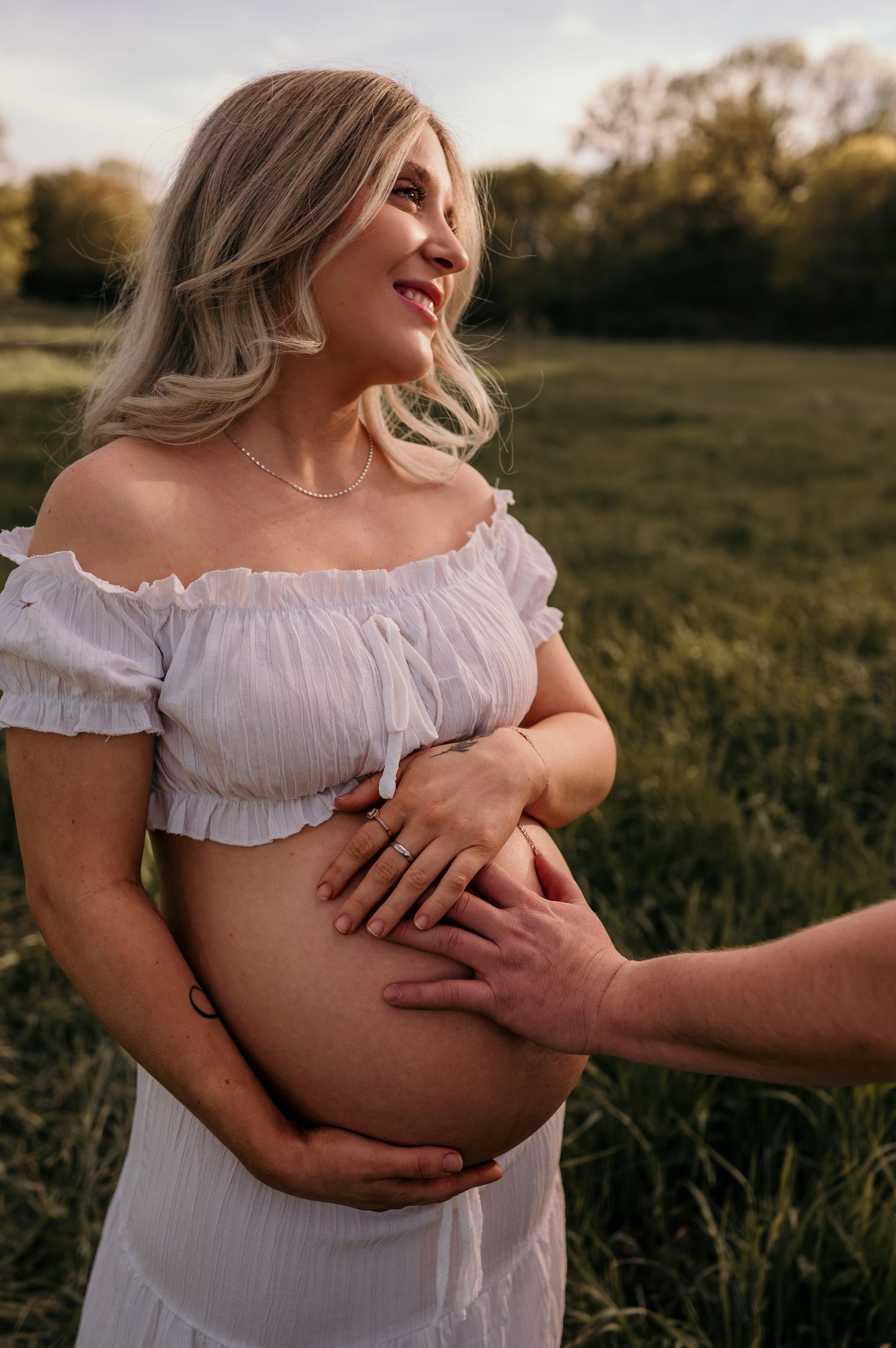 Smiling pregnant woman in white off-shoulder dress cradling her baby bump outdoors in a green field at golden hour.