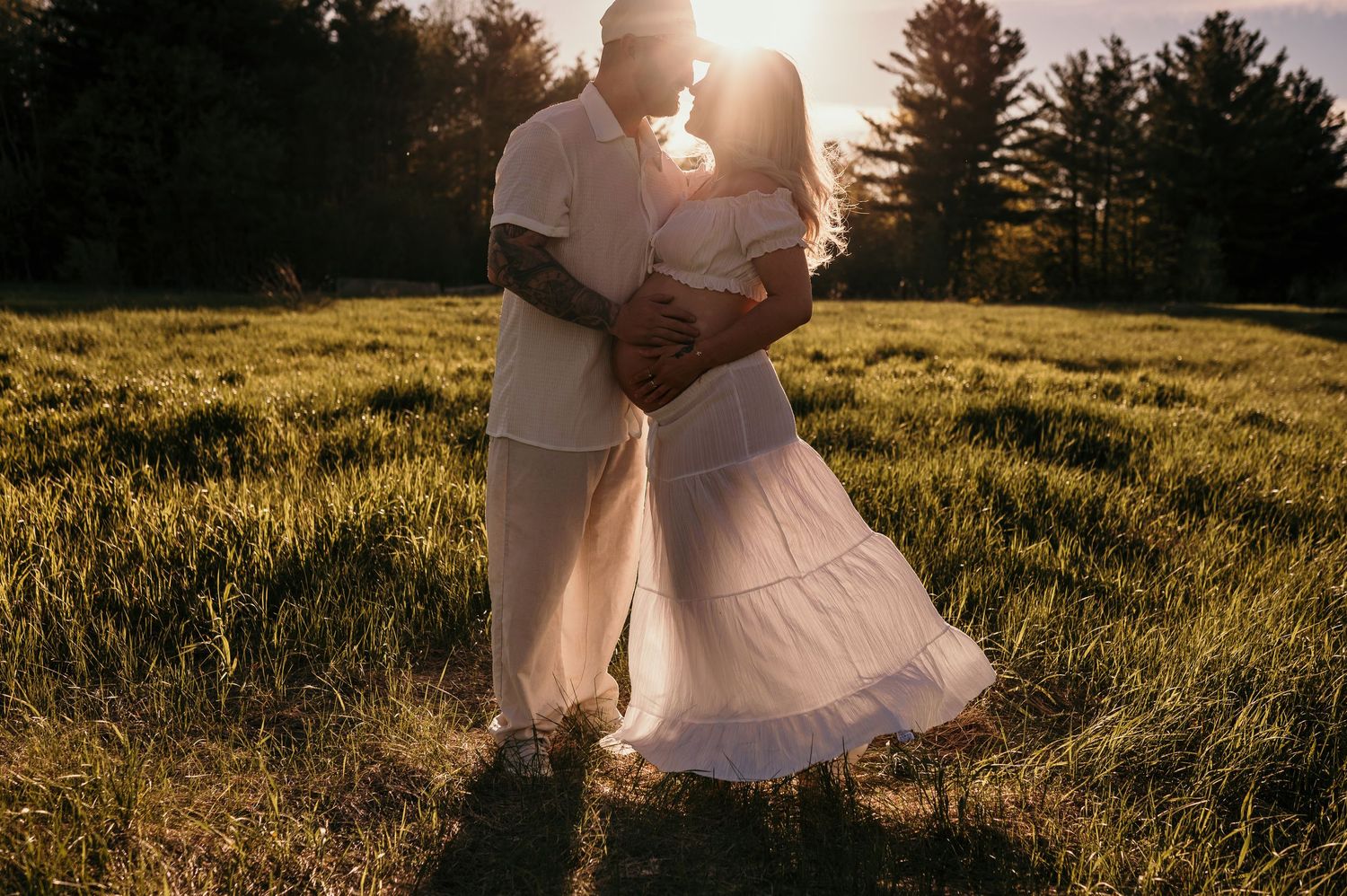 Couple embracing in golden sunset light in a lush green field, wearing white outfits, with trees in the background.