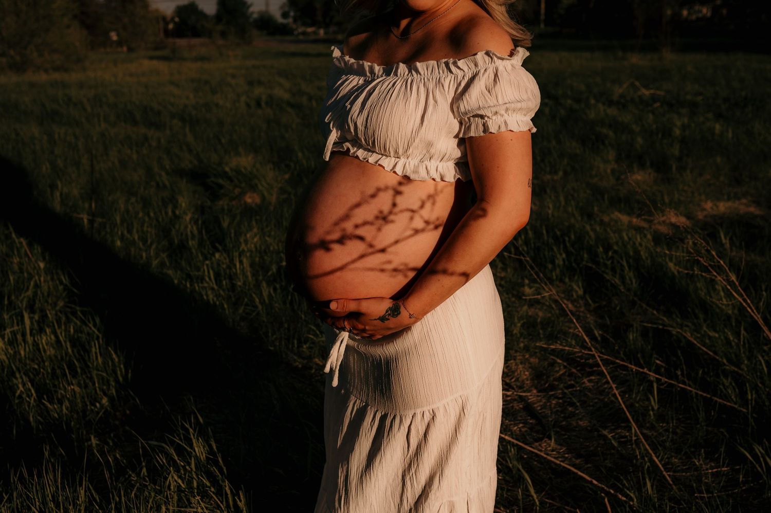 Pregnant woman in white dress cradling baby bump outdoors at golden hour in a lush green field.