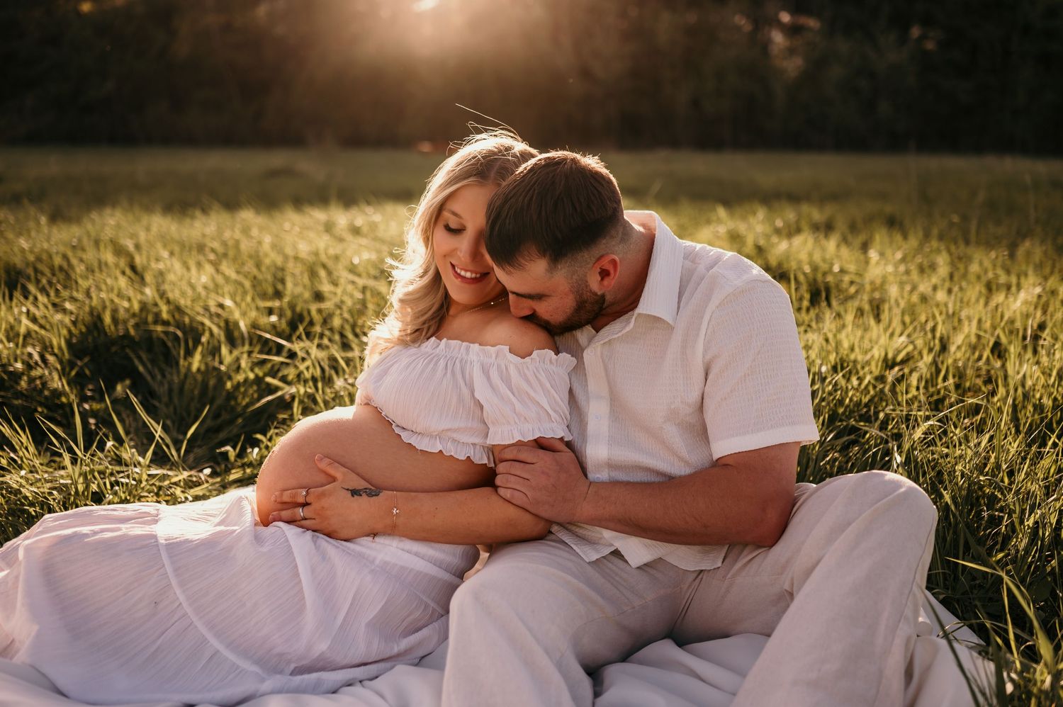 Couple sitting in sunlit field, man tenderly kissing pregnant woman's head, golden hour maternity photo.