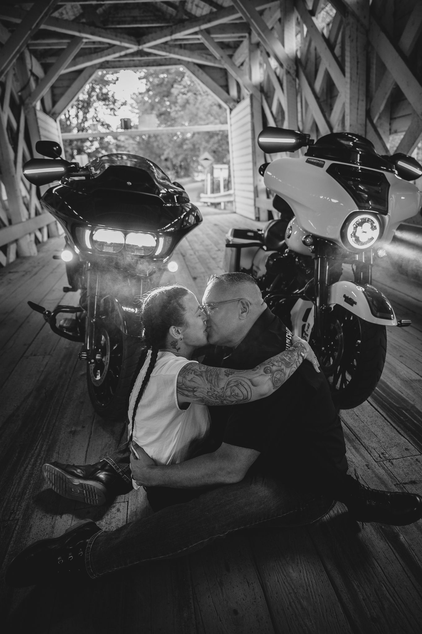 A black and white photo of two motorcycles parked inside a covered wooden bridge with figures sitting on the floor.