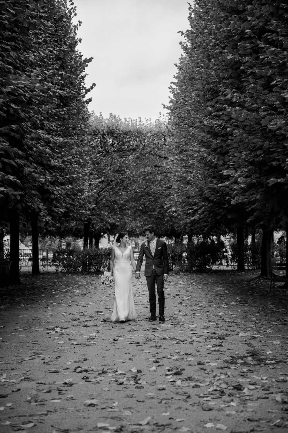Black and white photo of a bride and groom walking hand in hand through a tree-lined path scattered with fallen leaves.