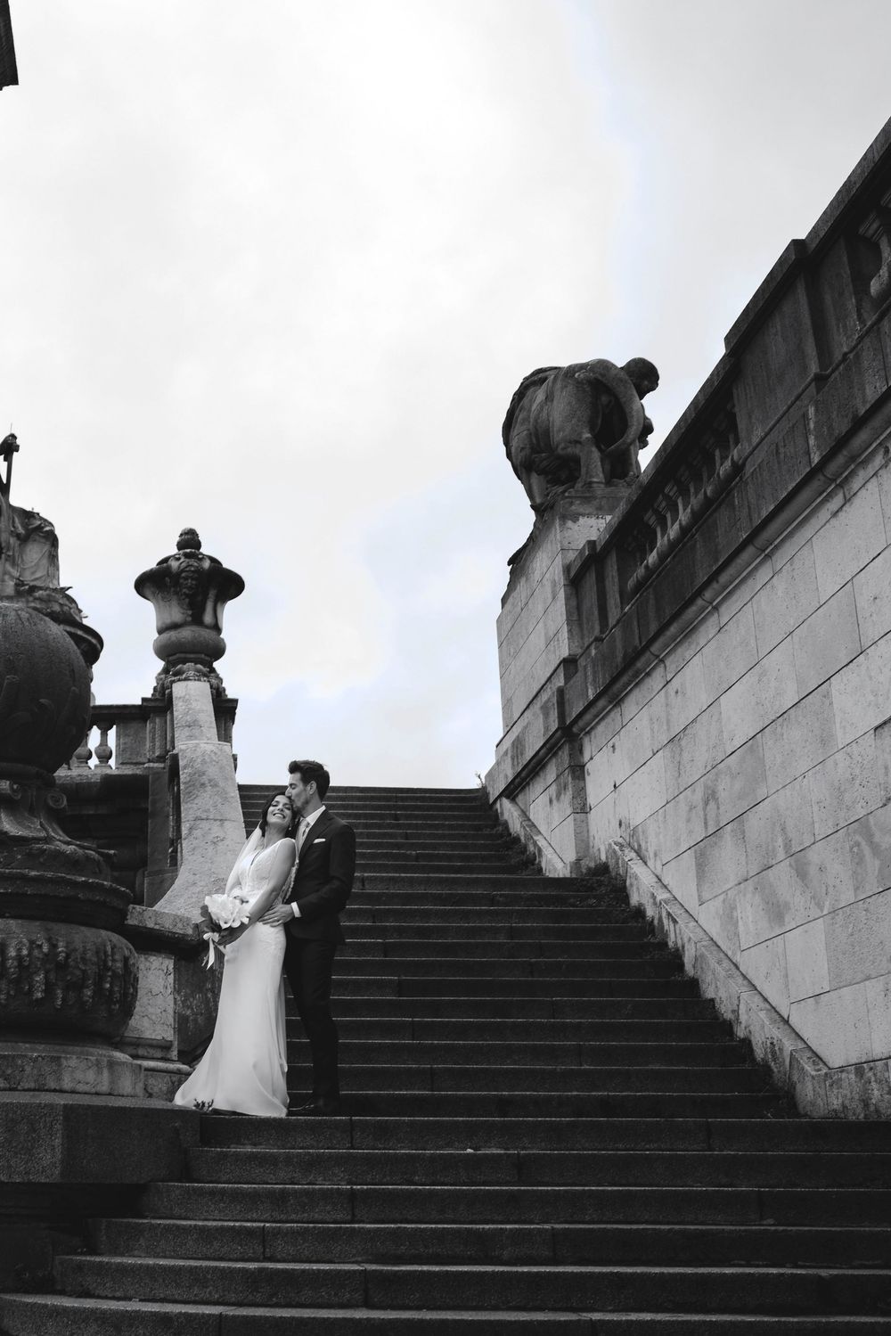 Black and white photo of a bride and groom posing on grand stone steps with classical sculptures.