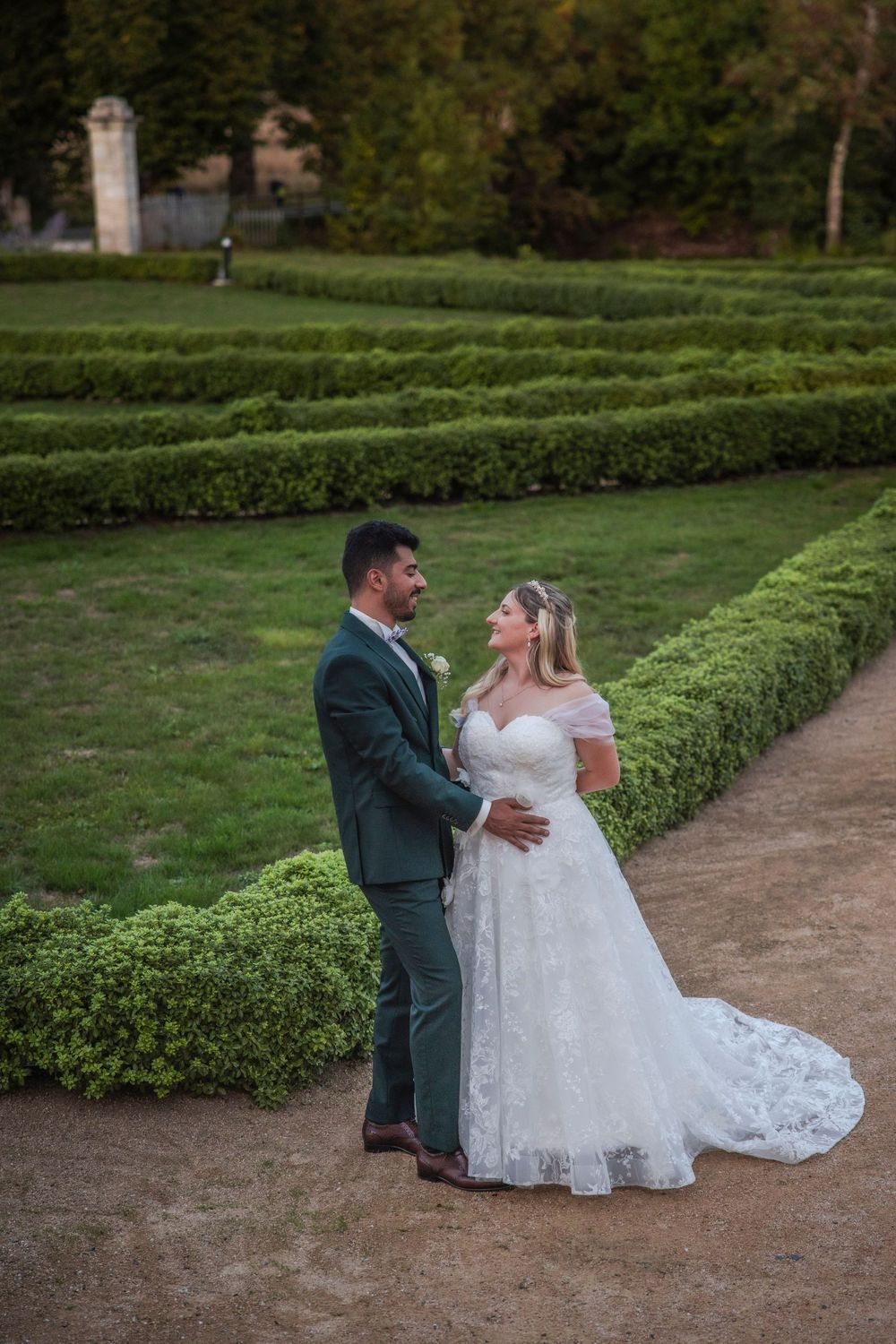 A couple in wedding attire poses together on a garden path surrounded by manicured hedges.