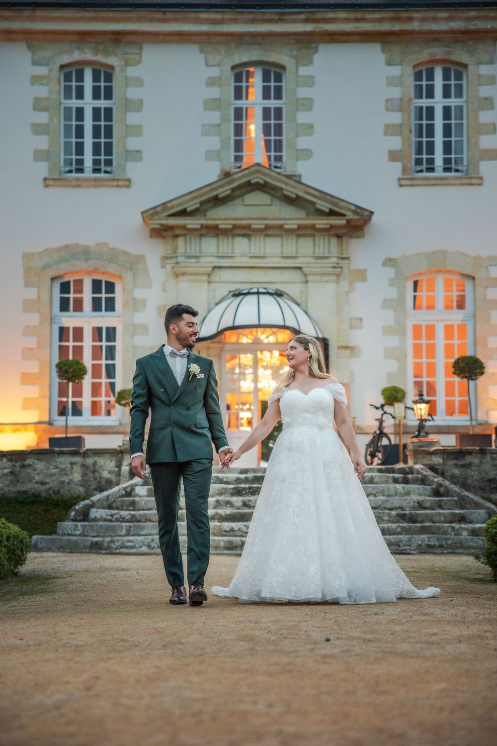 A couple in wedding attire stands on stone steps in front of an elegant illuminated French chateau at dusk.