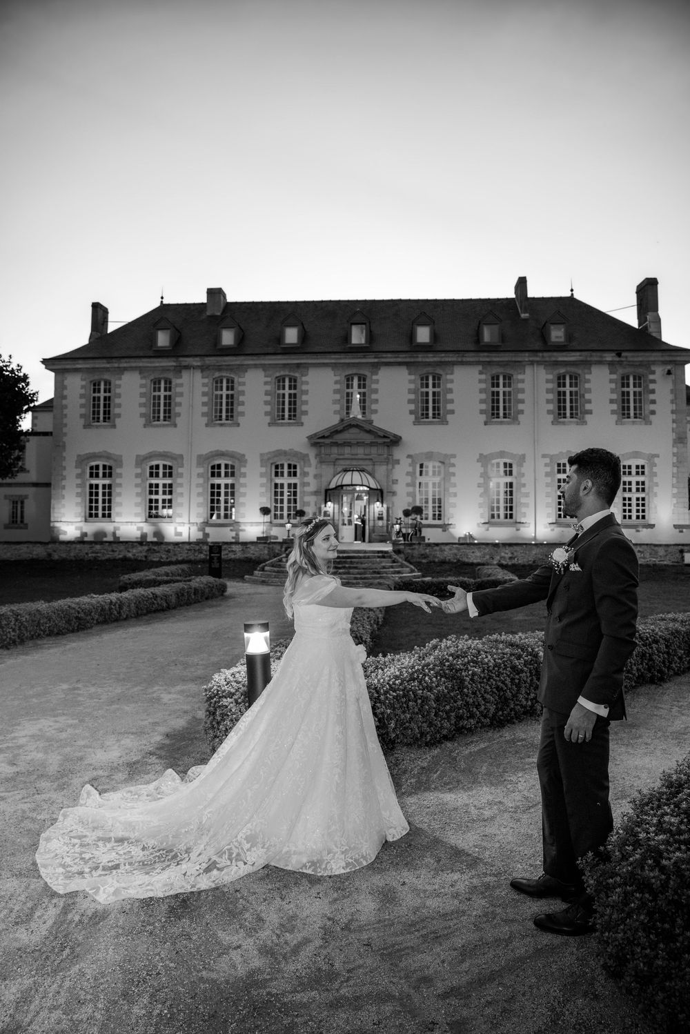 A couple in wedding attire poses together in front of a grand historic mansion with dramatic black and white photography.
