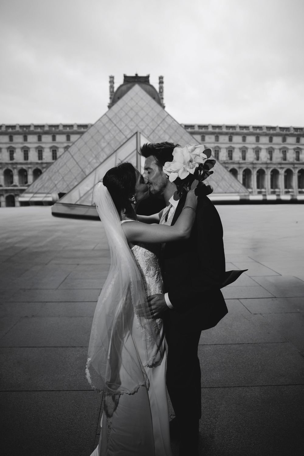 Bride and groom kissing in front of the Louvre Pyramid in Paris in a black and white wedding photo.